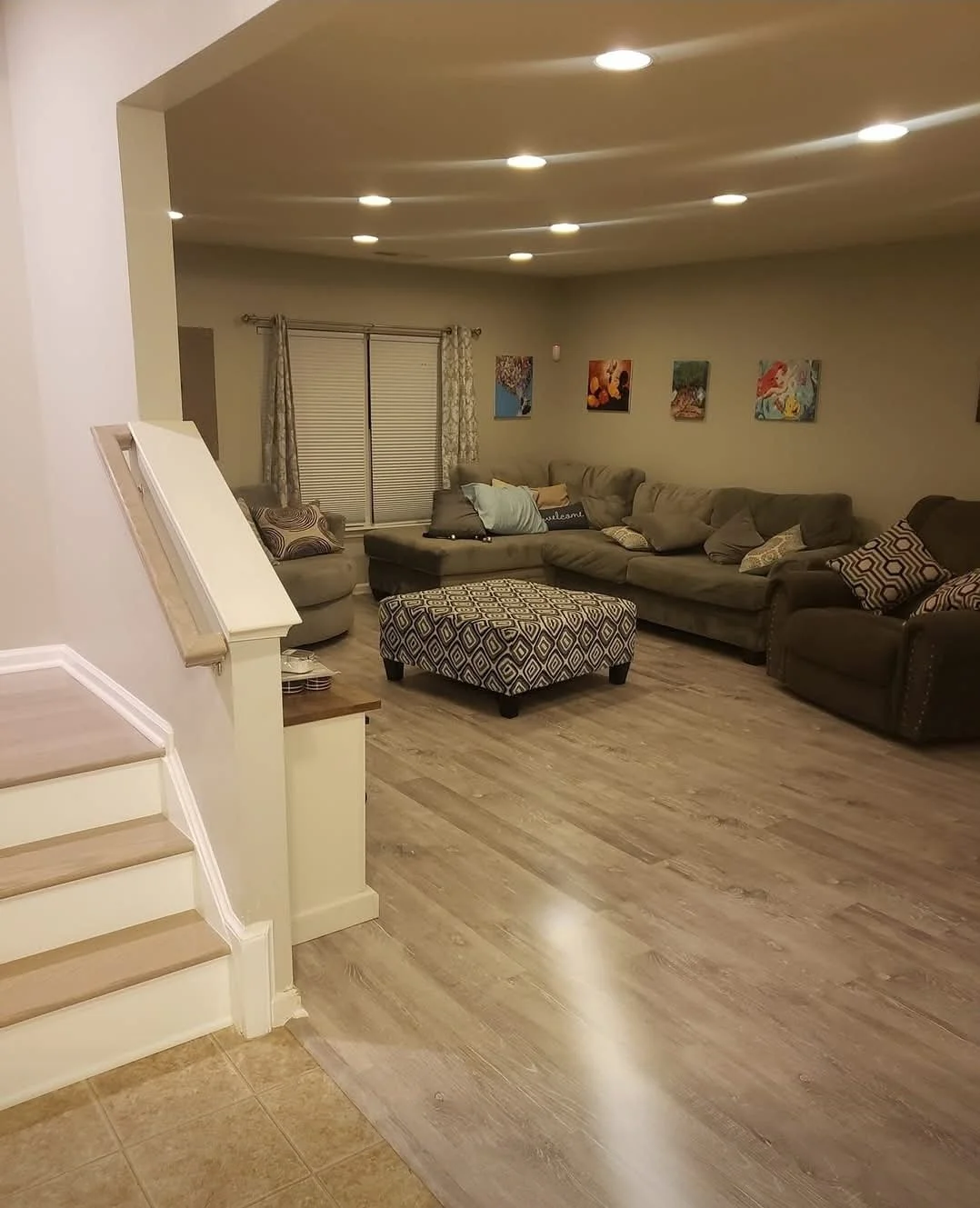 A cozy living room with a large sectional sofa, patterned ottoman, and colorful wall art, illuminated by ceiling recessed lights.