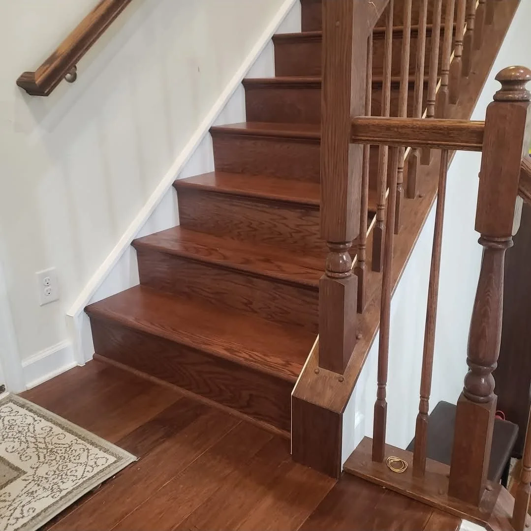 Wooden staircase with handrail and balusters, carpeted area with a decorative mat, and an electrical outlet on the wall.
