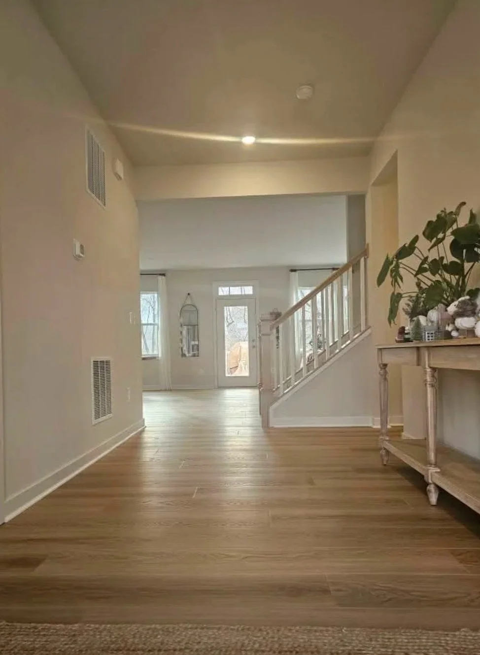 A spacious living room with light-colored walls, hardwood flooring, a front door, large windows, a staircase with white railing, a small side table, and a large potted plant.