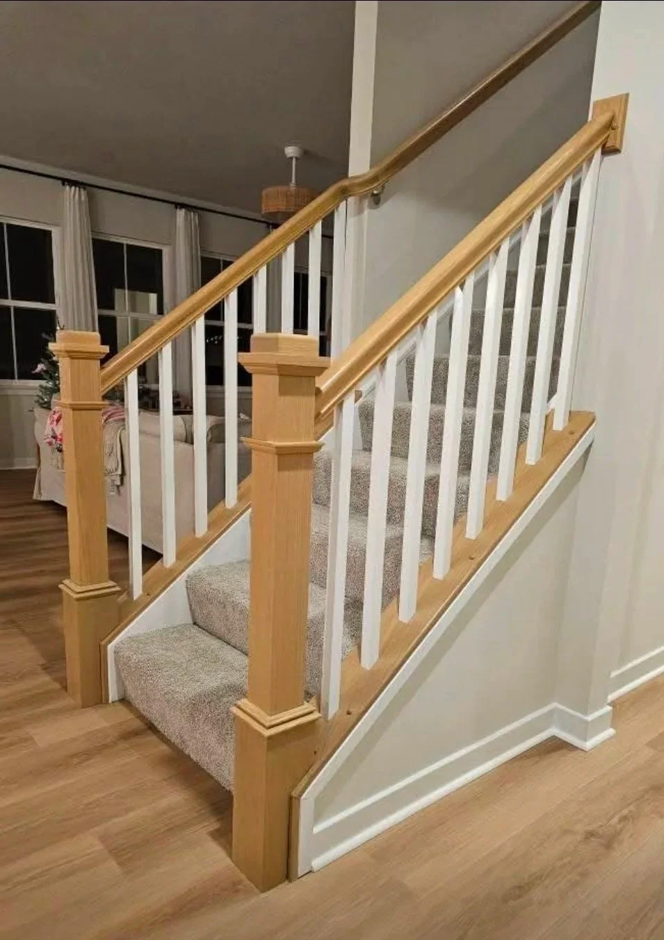Staircase with carpeted steps, wooden handrails, and white spindles. Part of a living room with a sofa and large windows in the background.
