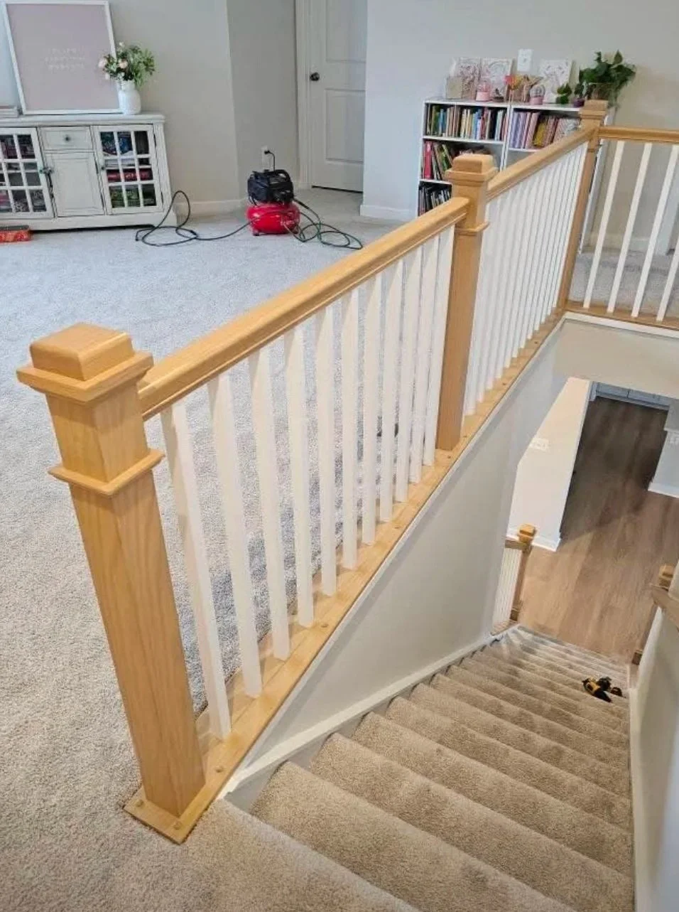 The image shows a staircase with a wooden and white railing overlooking a carpeted upper level. On the upper level, there is a white media console with a potted plant and some decorative items. A bookshelf filled with books and decorative pieces is v