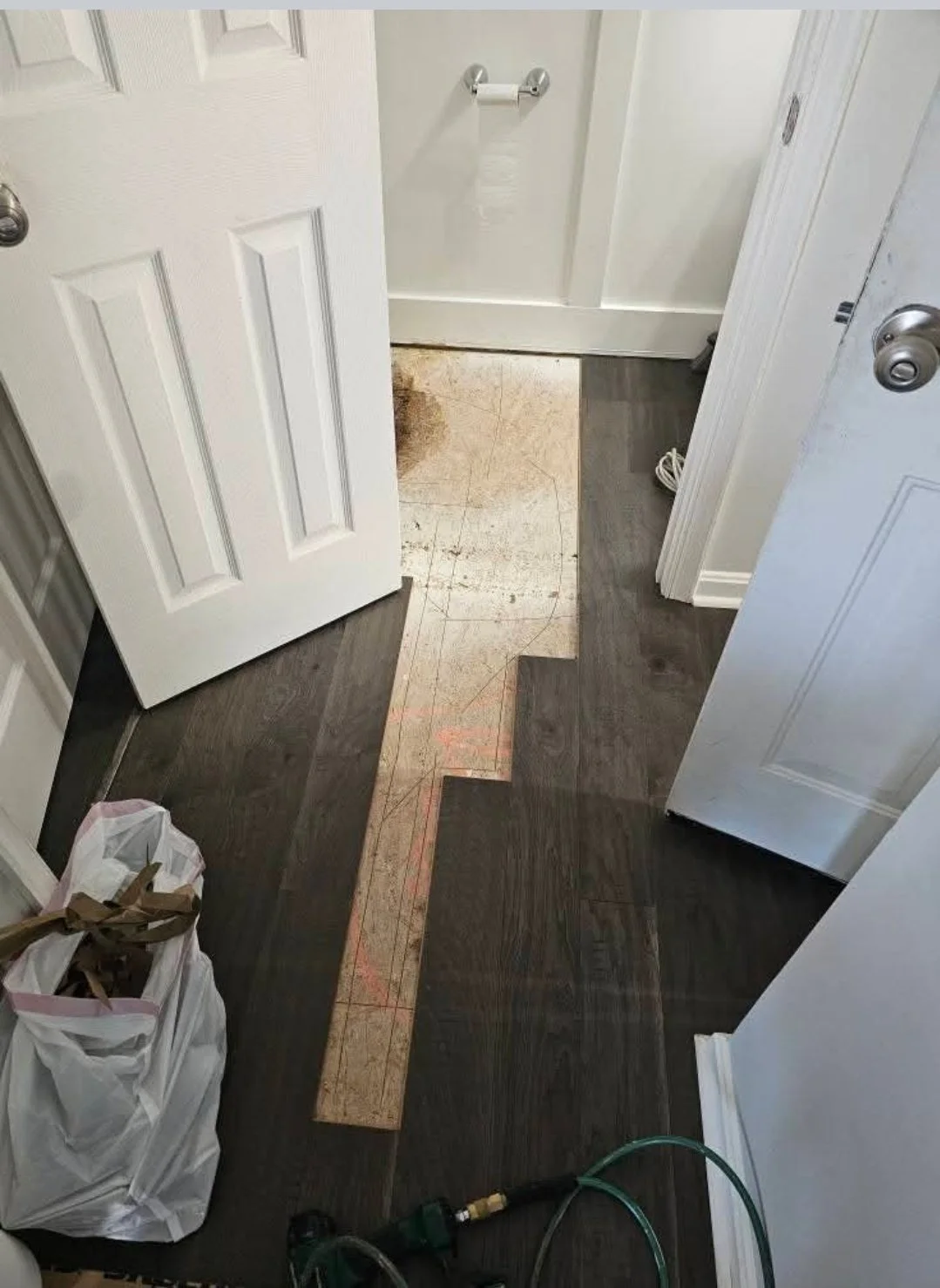 This image shows a section of a room, likely a bathroom, with a partially removed floor revealing a plywood subfloor underneath. The flooring is dark wood, and some tools and a trash bag are present, indicating ongoing flooring work.