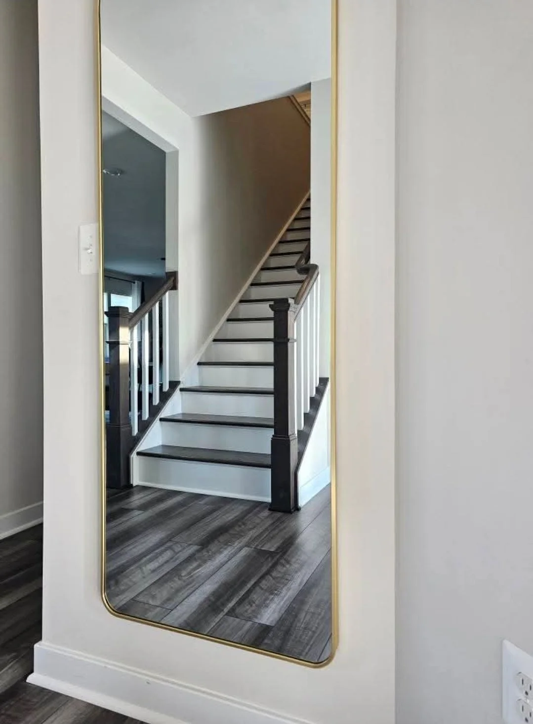 A tall mirror with a gold border reflecting a staircase with dark wood steps and white risers, and a wooden handrail with black posts.