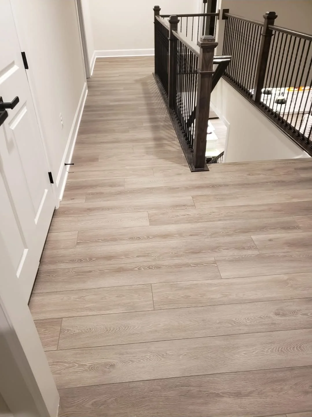 Interior view of a house showing a staircase with black wooden railing, hardwood flooring, closed white door, and beige walls.