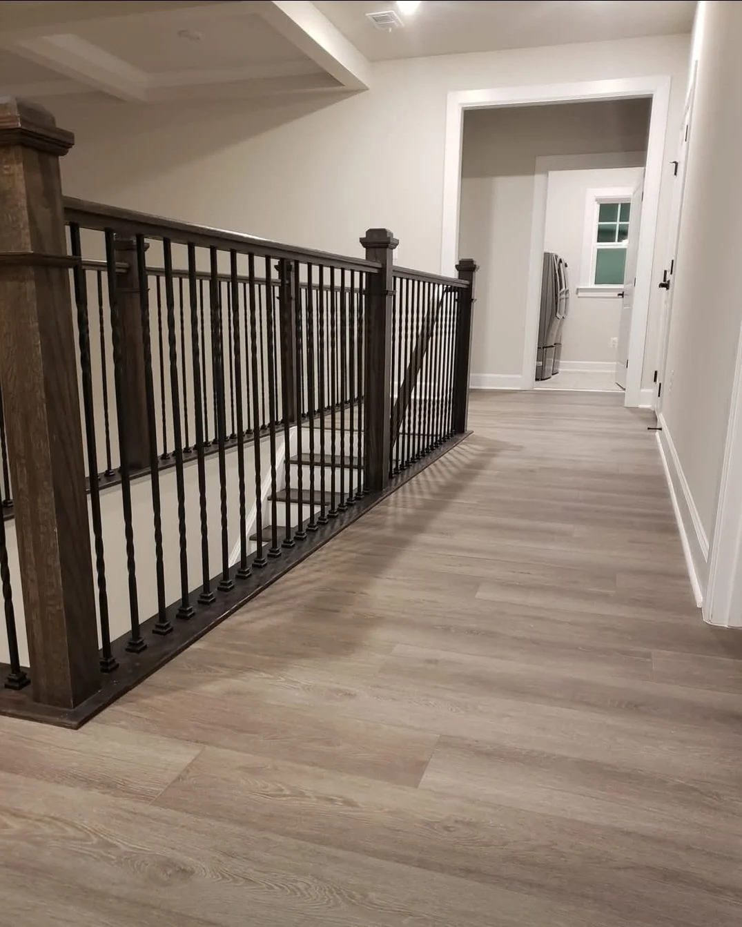 Interior view of a modern home hallway with a wooden railing and stairs on the left, light-colored wood flooring, and a laundry area with a washing machine visible through an open doorway at the end.
