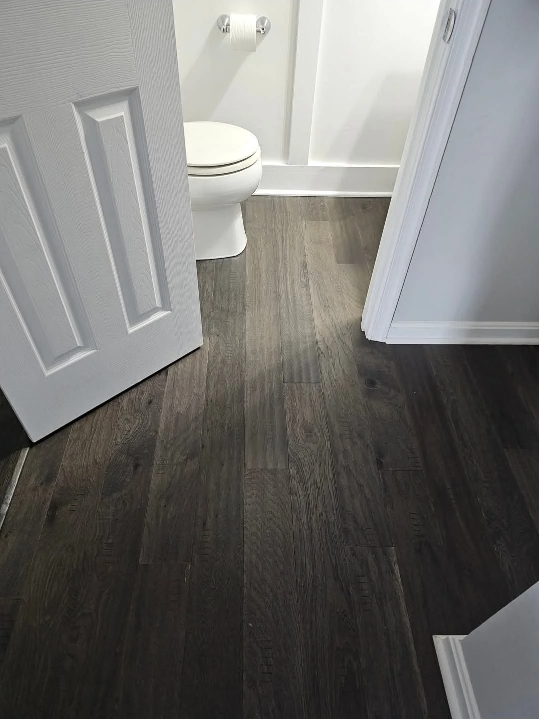 View of a bathroom with a white toilet, white walls, wood-look flooring, and a partially open door.