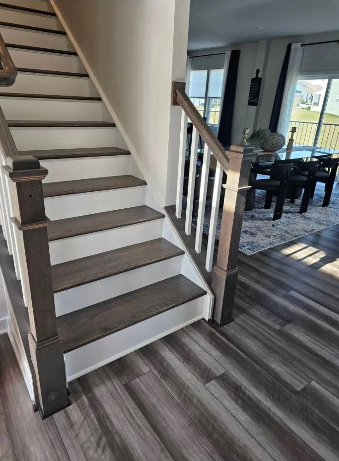 Interior view of a staircase with dark wood steps and white risers, next to a living or dining area with hardwood flooring, a large window, and a glass door leading outside.