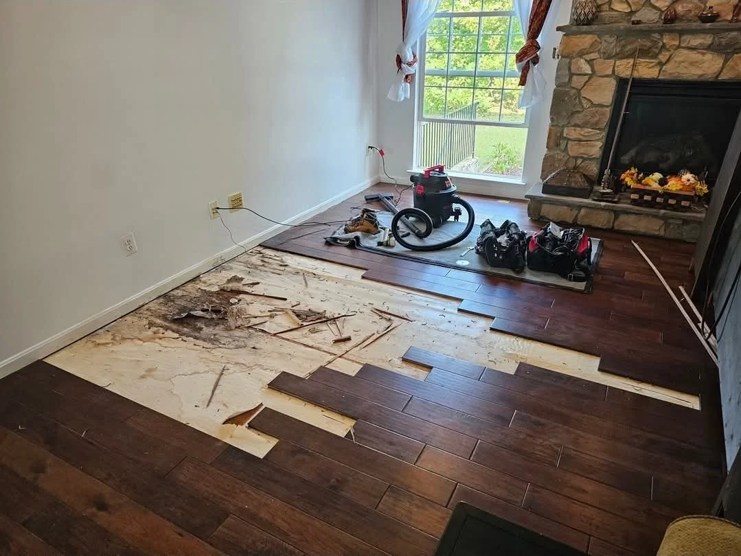 Living room with partially removed hardwood flooring, exposing subfloor. Tools and equipment for flooring installation are on the floor, with a large window and stone fireplace visible.