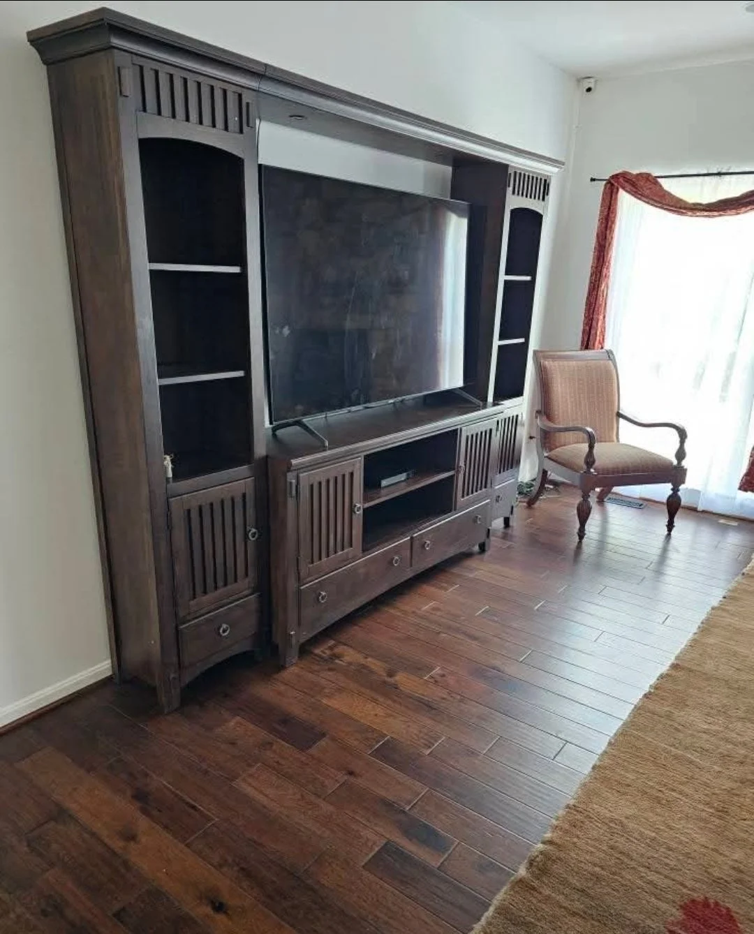 A television entertainment center with open shelves and drawers, situated on a wooden floor near a window with curtains. A single wooden armchair is placed beside it.