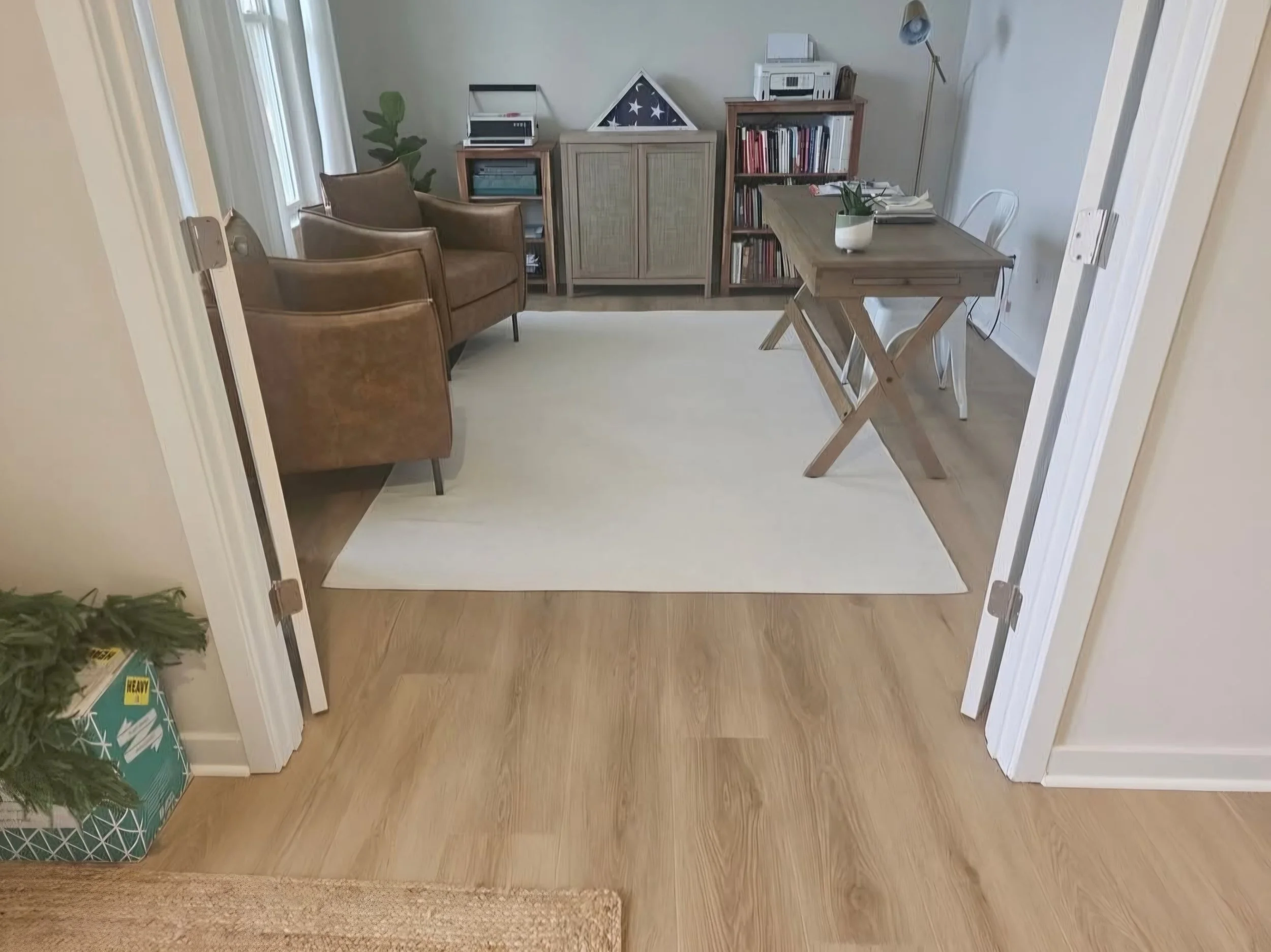 View of a living room with two brown chairs, a wooden desk with a white chair, a bookshelf, a cabinet with a flag display, a lamp, a potted plant, and a lamp standing in the corner. The room has a light-colored carpet and wood flooring, seen through 