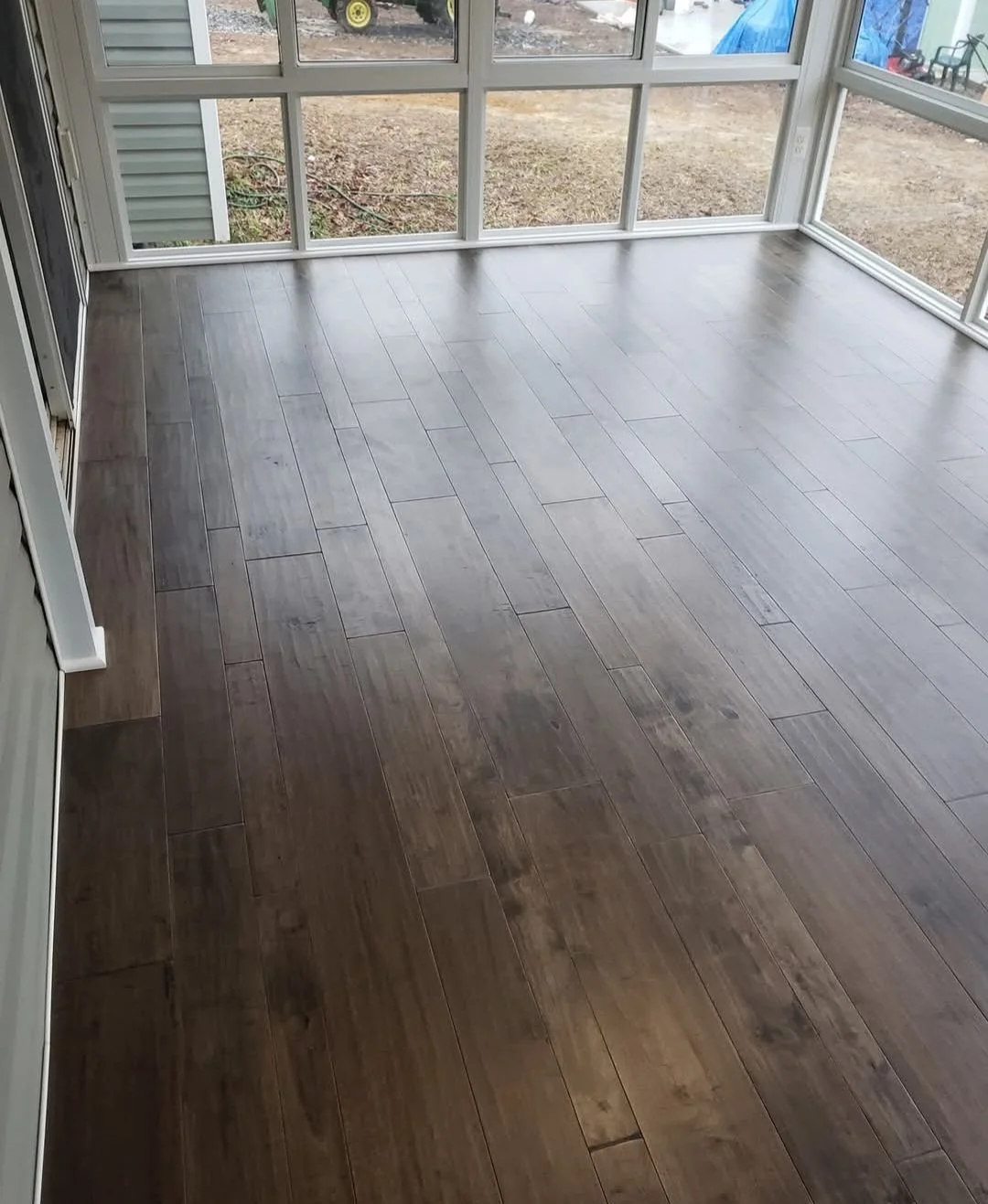 A room with wooden flooring and large windows showing an outdoor area with dirt ground, some equipment, and a blue tarp.