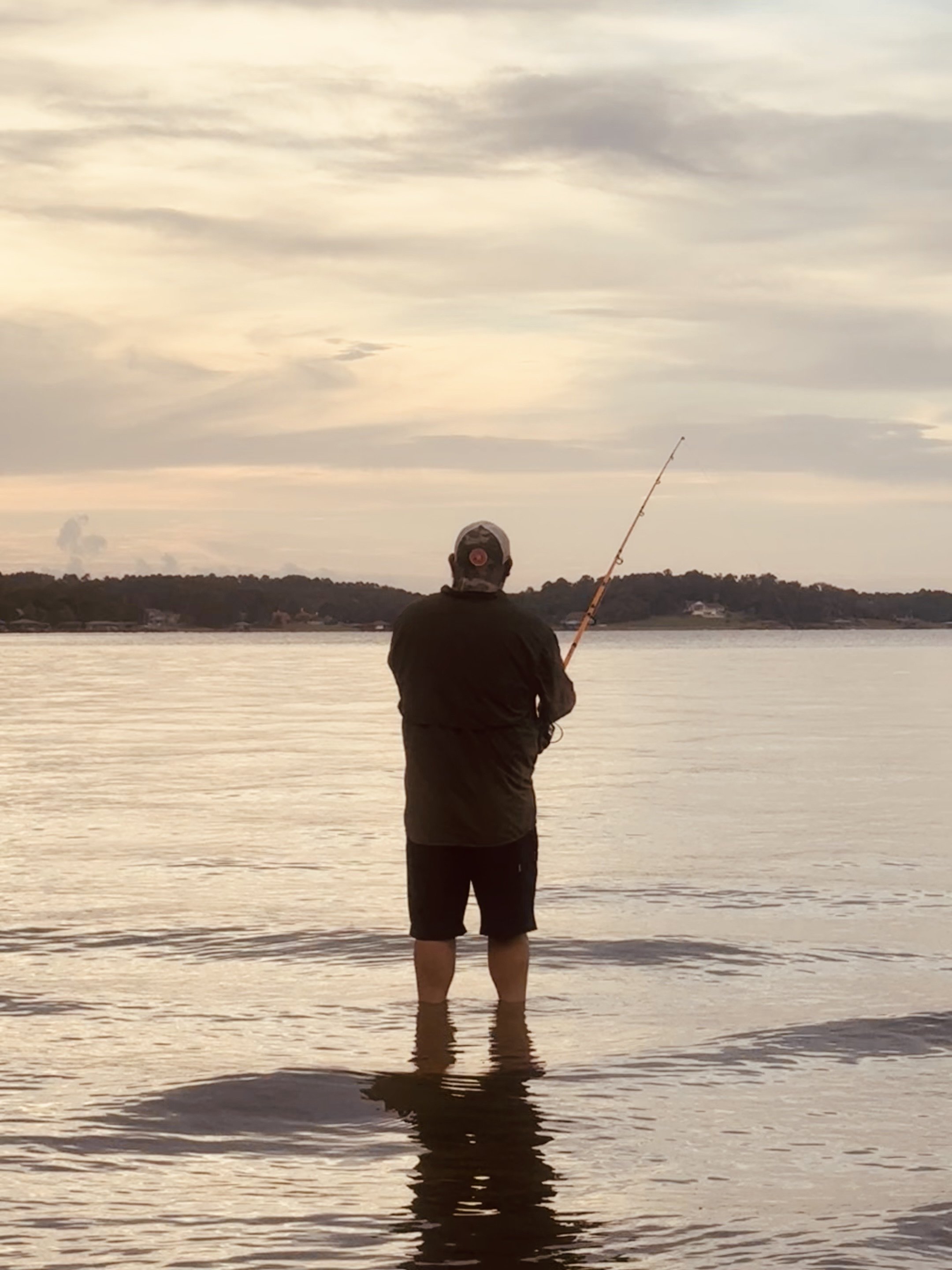 A person fishing in a calm body of water during sunset or sunrise, standing with their back to the camera and holding a fishing rod.