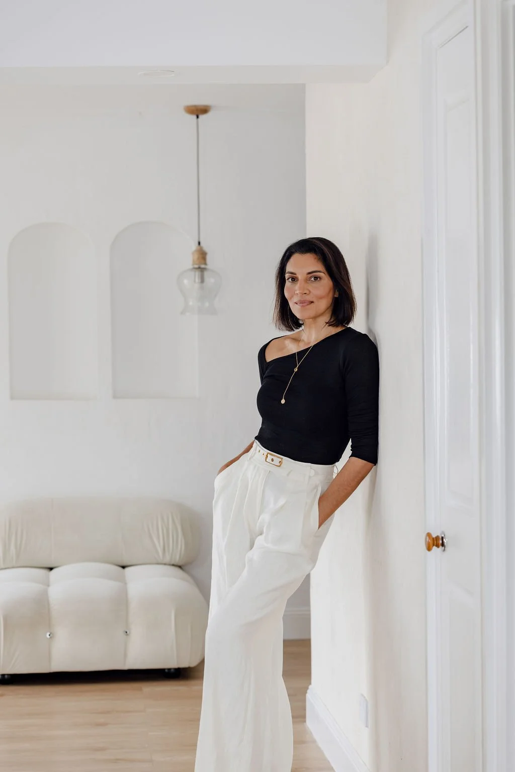 A woman with shoulder-length dark hair, wearing a black top and white high-waisted trousers, stands in a bright, minimalistic living room with white walls and a beige sofa.
