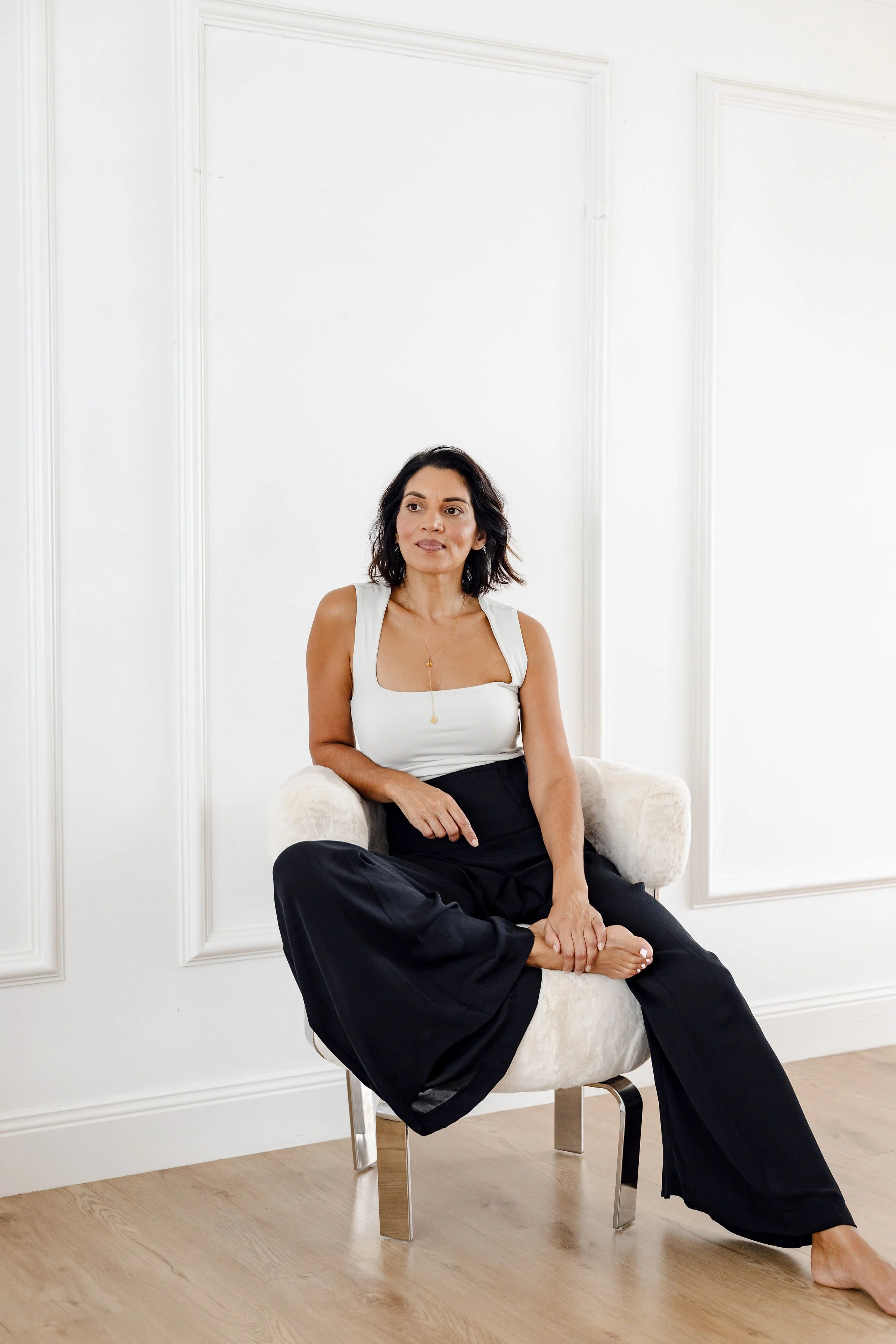 A woman sits on a light-colored chair in a room with white paneled walls and wooden flooring. She has shoulder-length dark hair and is wearing a white sleeveless top and black wide-leg pants.