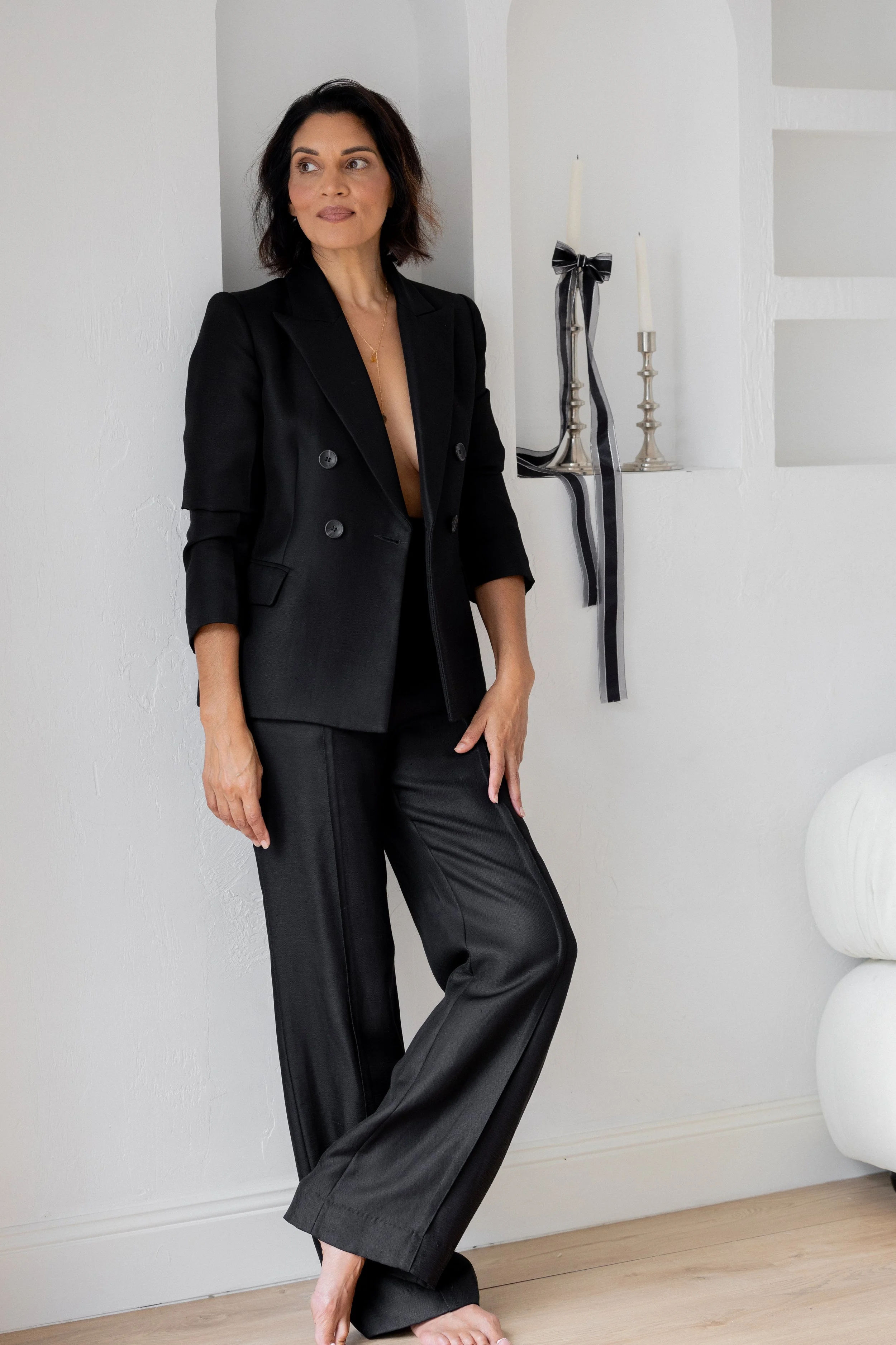 Woman with shoulder-length dark hair wearing a black blazer and black leather pants, standing barefoot in a white room with candles on a shelf.