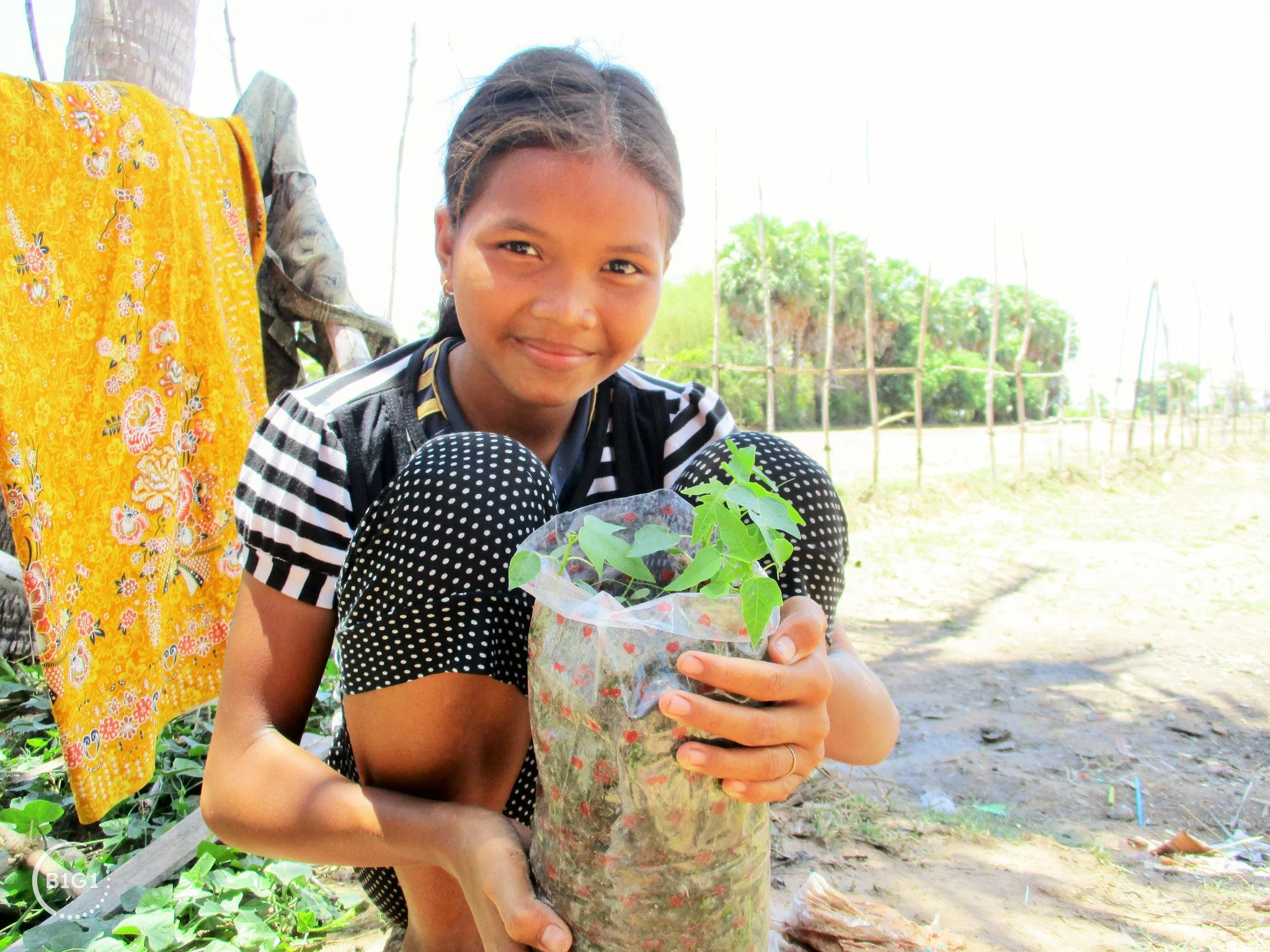A young girl smiling and holding a small potted plant outside in a rural area.