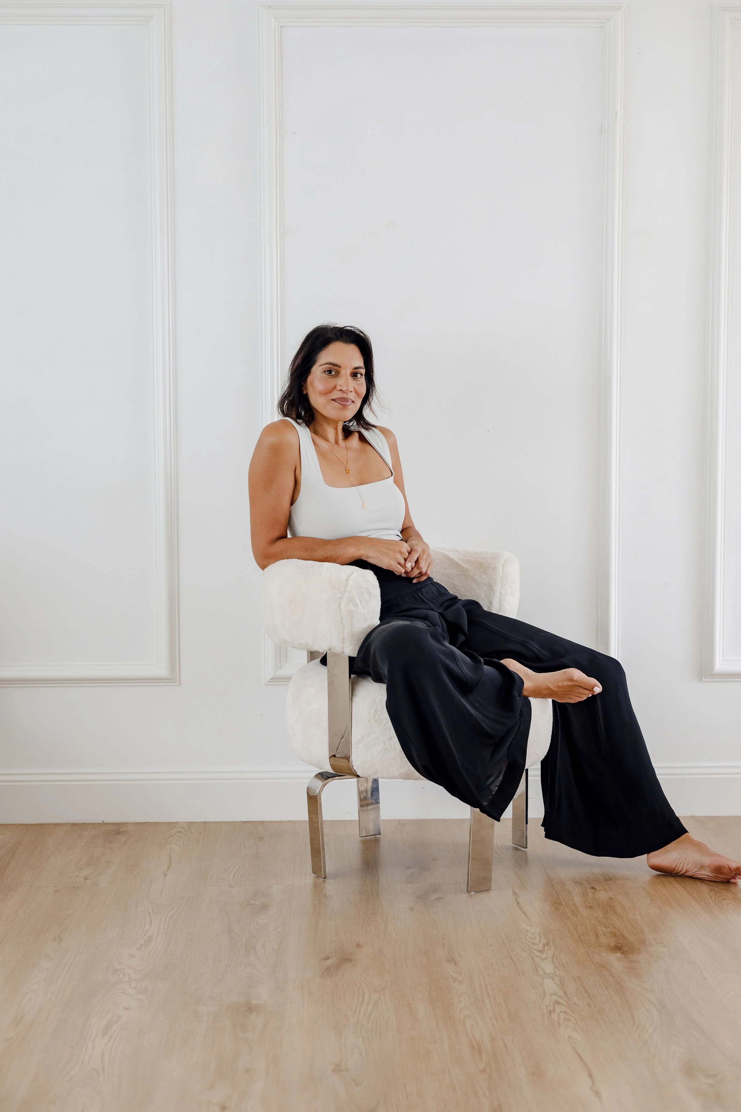 A woman with dark hair in a white sleeveless top and black wide-leg pants, sitting barefoot on a white, plush, modern chair with metal legs, in a room with white walls and light wood flooring.