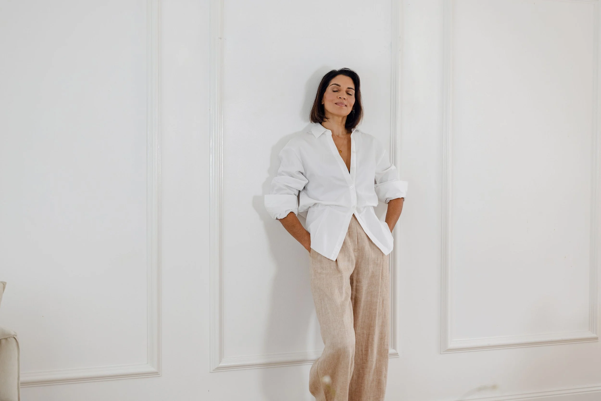 Woman standing against a white wall with her hands in her pockets, wearing a white button-up shirt and beige pants, with a relaxed expression.