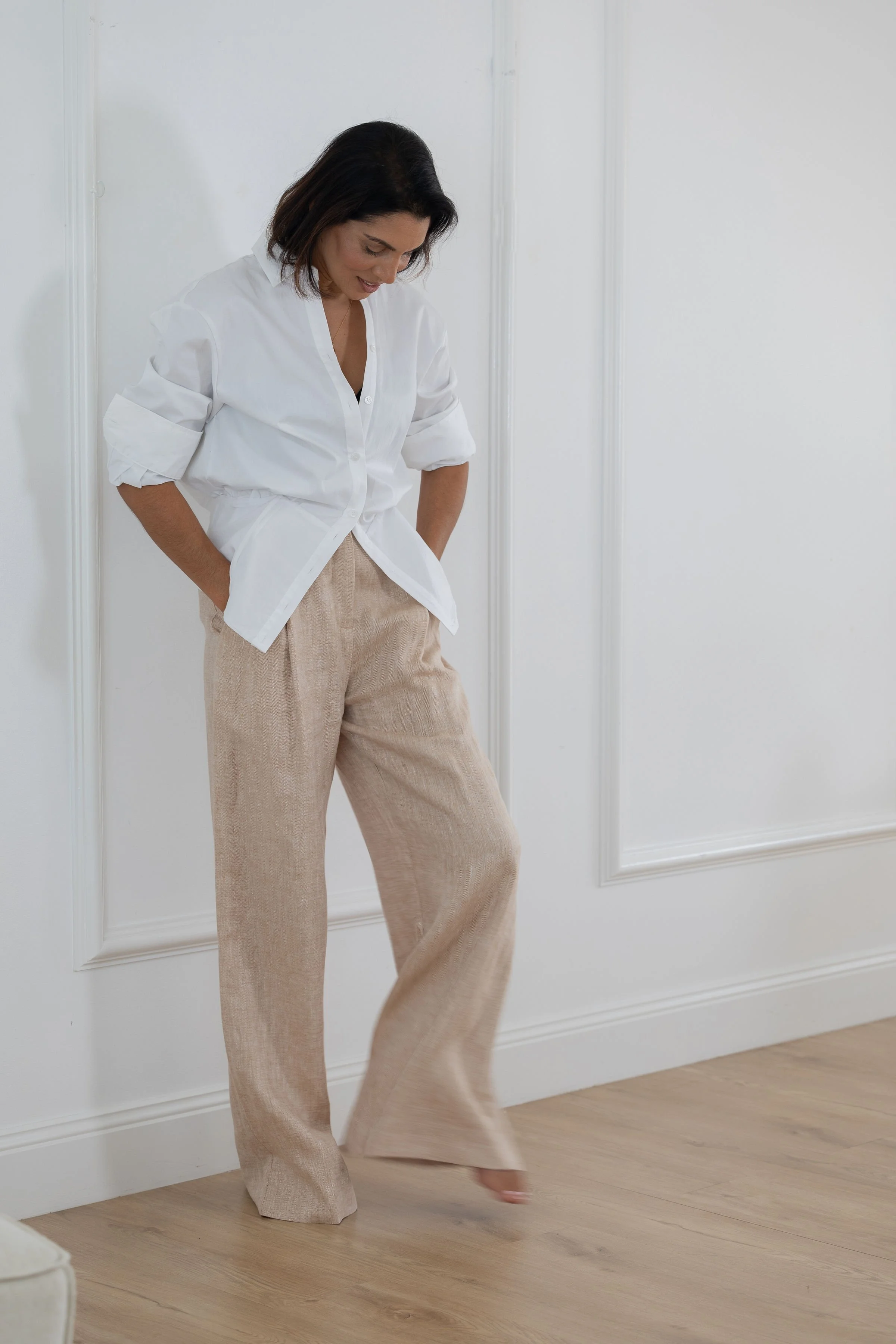 Woman standing in a white room, looking down with hands in her beige linen pants pockets, wearing a white button-up shirt with rolled-up sleeves.