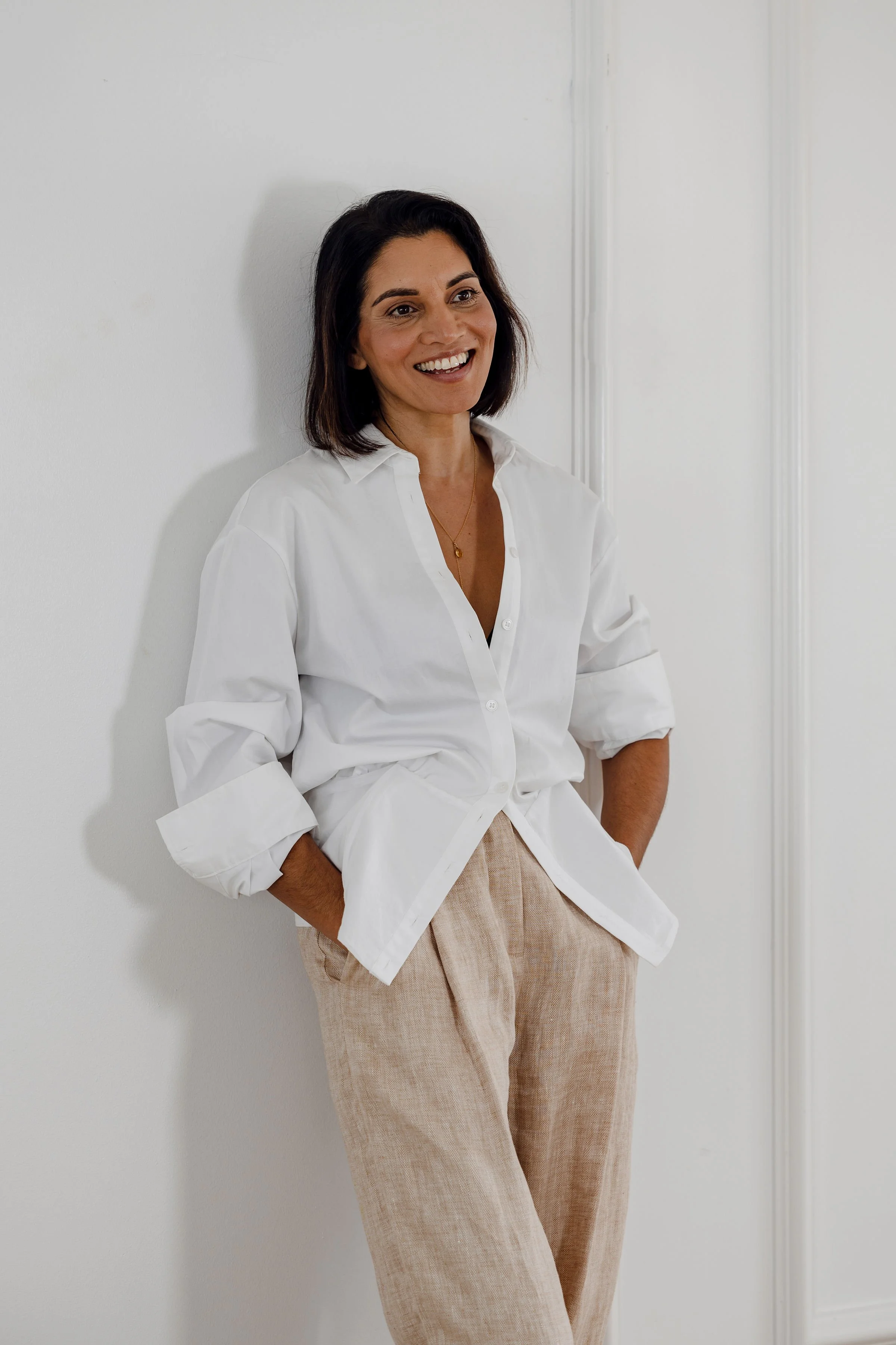 A woman with shoulder-length dark hair smiling while leaning against a white wall, wearing a white button-up shirt with rolled sleeves and beige pants.