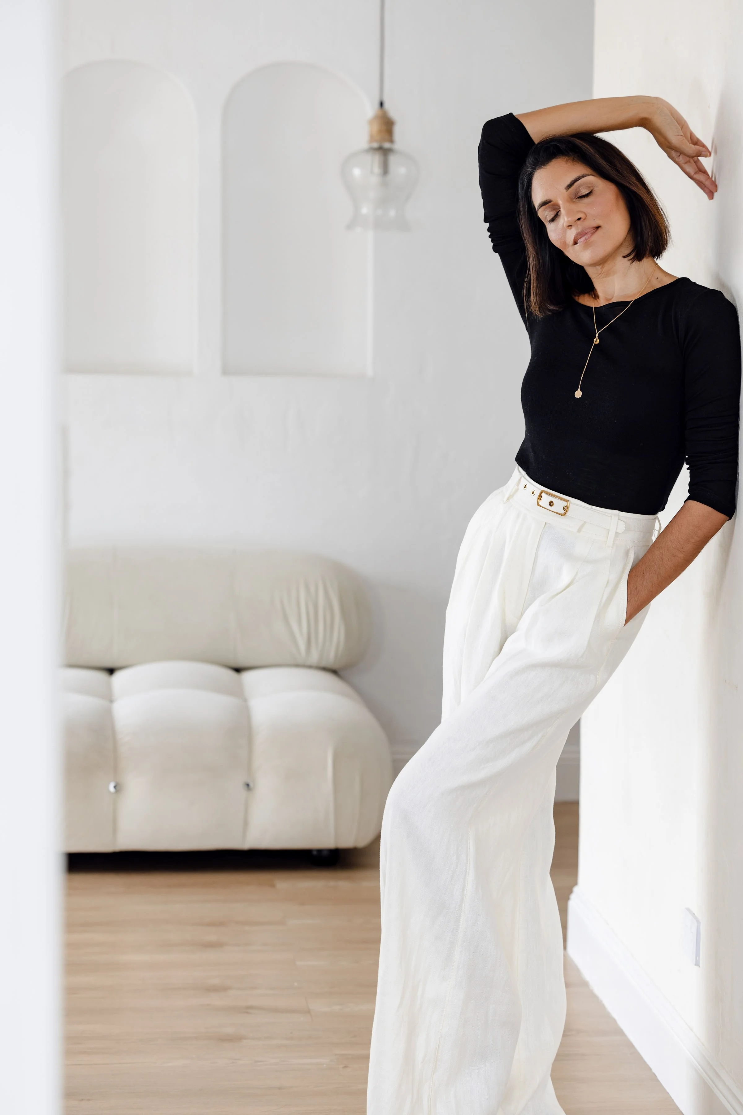 A woman standing against a white wall with her eyes closed, wearing a black top and white wide-leg pants in a minimalist room with wooden flooring and a beige sofa.