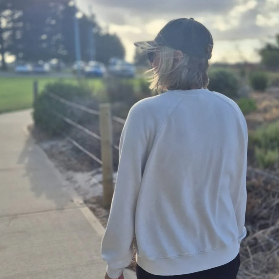 A person with shoulder-length hair wearing a black baseball cap, sunglasses, a white oversized sweatshirt, and dark pants, walking along a sidewalk at sunset with clouds in the sky and cars in the background.