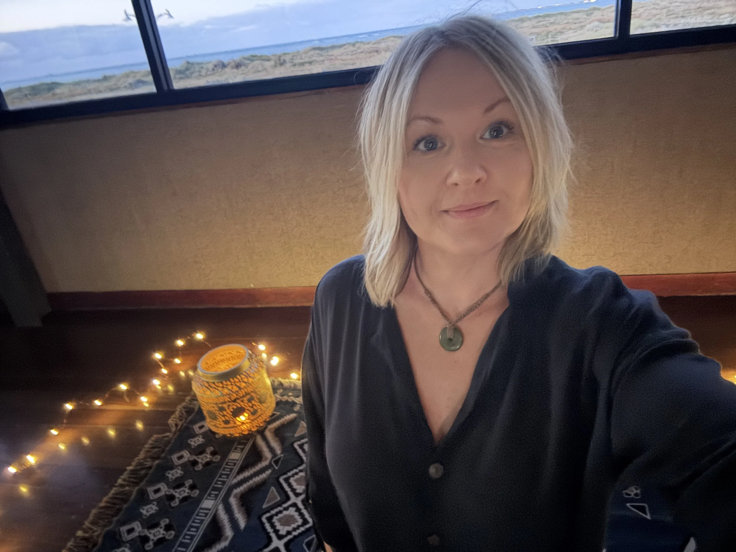 A woman with blond hair and blue eyes taking a selfie at a yacht club with a view of the ocean and sky in the background. The interior includes string lights and patterned decorations.