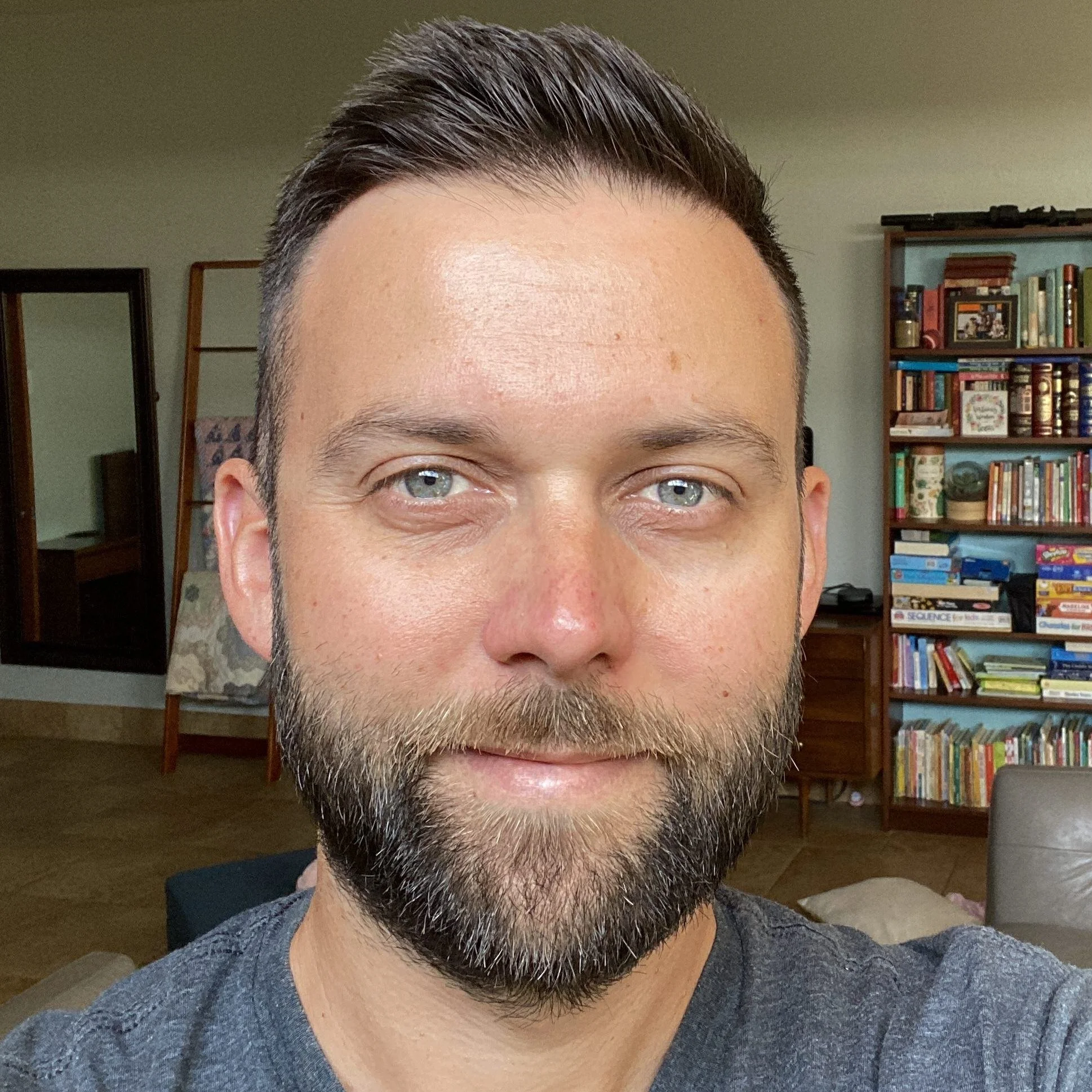 Close-up of a man with short dark hair and a beard, smiling slightly, in a room with bookshelves and furniture in the background.