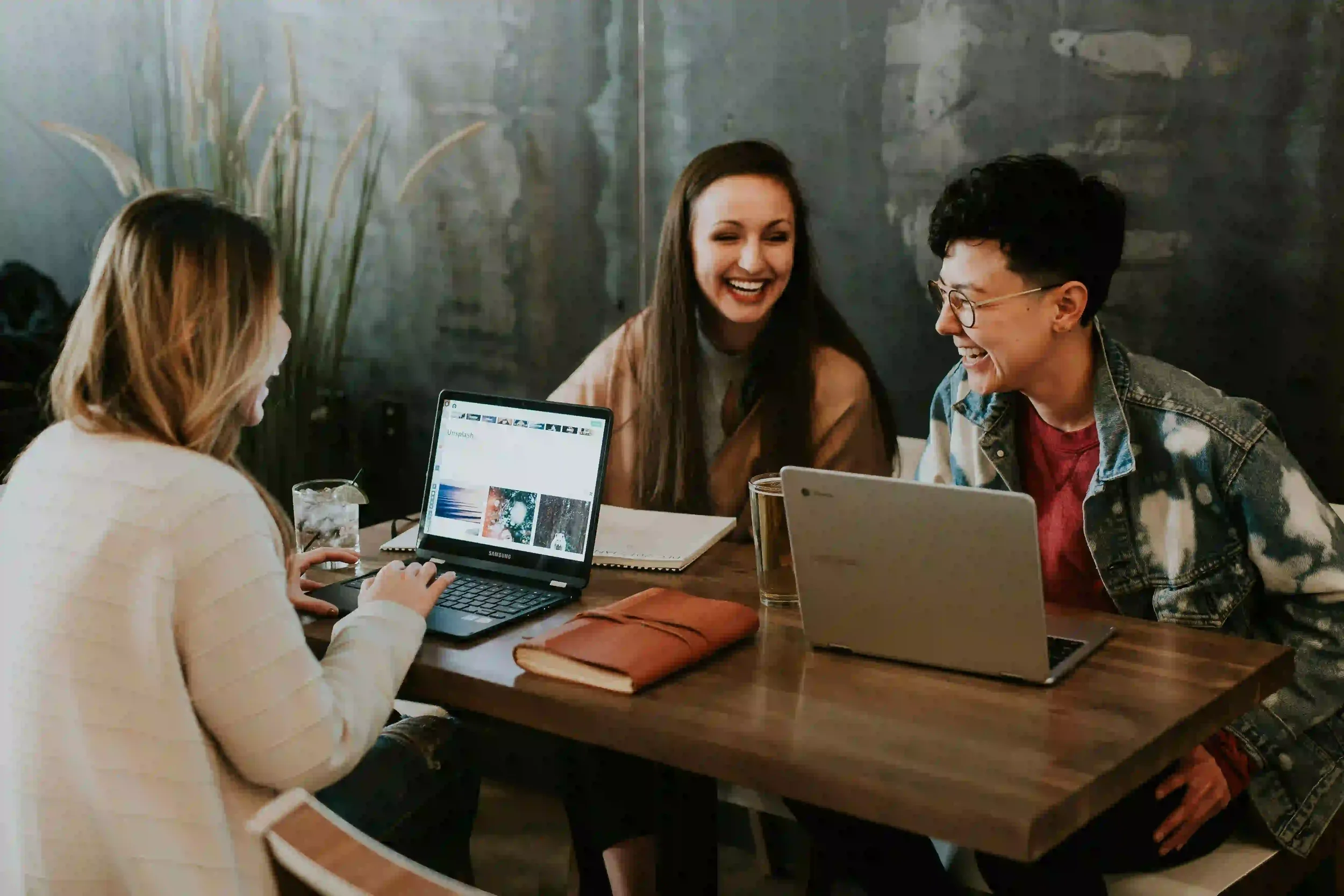 Three young women sitting at a wooden table, laughing and engaging in conversation. Two of them have laptops open, and there are notebooks and drinks on the table. The background features a dark, textured wall and some green plants.