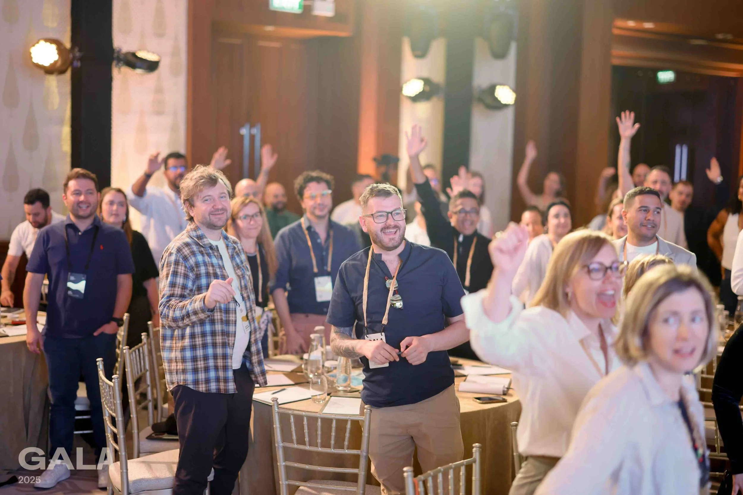 Group of smiling people at a conference or event, standing with some raising their hands in a large, well-lit room.