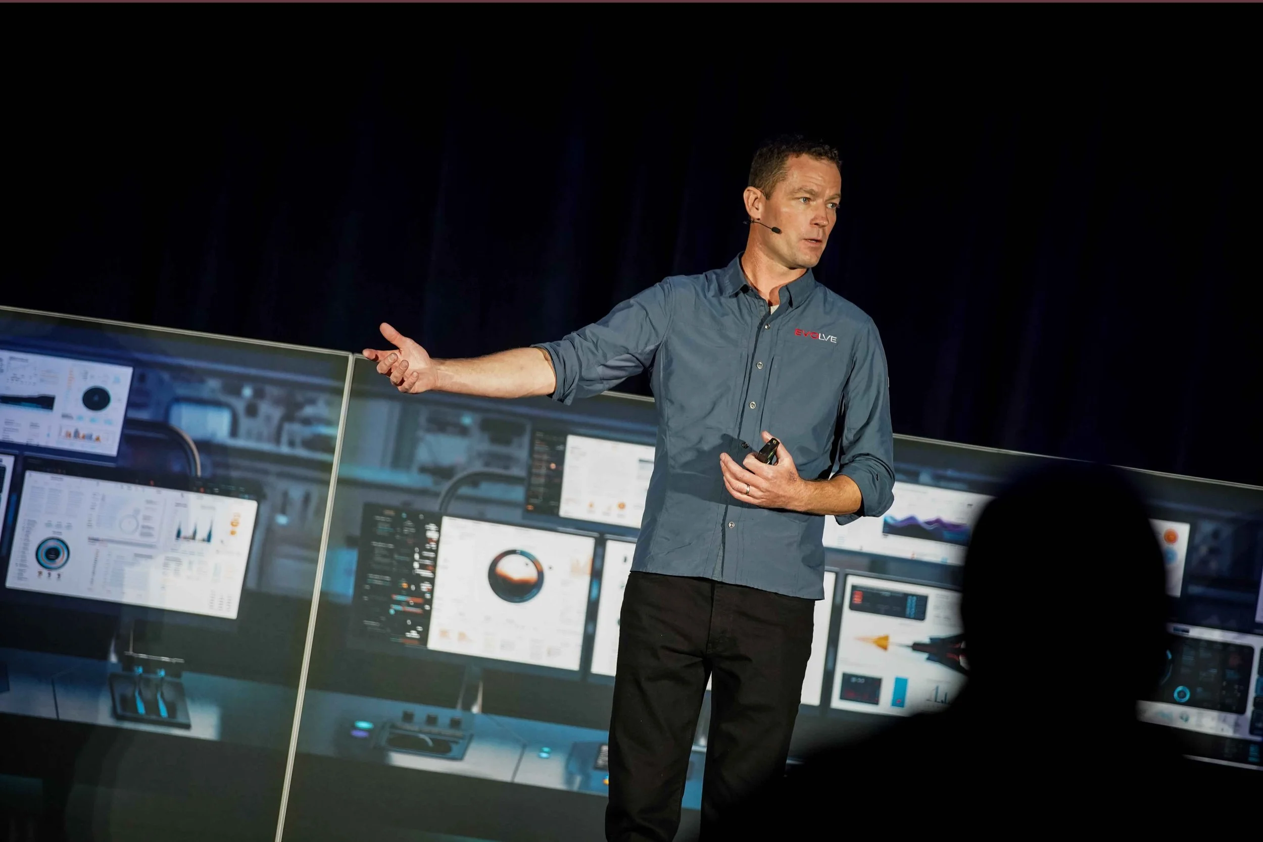 A man giving a presentation on a stage with multiple computer screens displaying graphs and data.