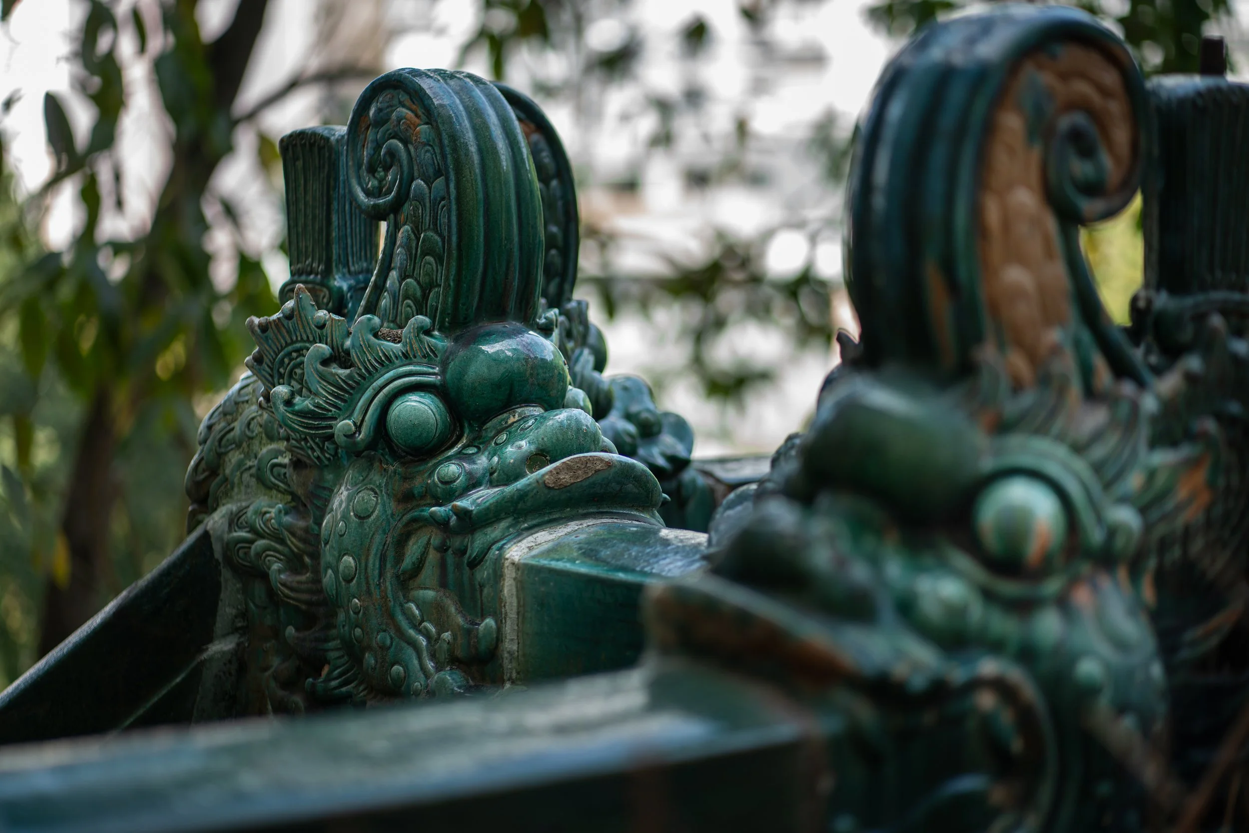 Close-up of ornate, green ceramic tiles with dragon motifs on a traditional architectural structure, with blurred greenery in the background.