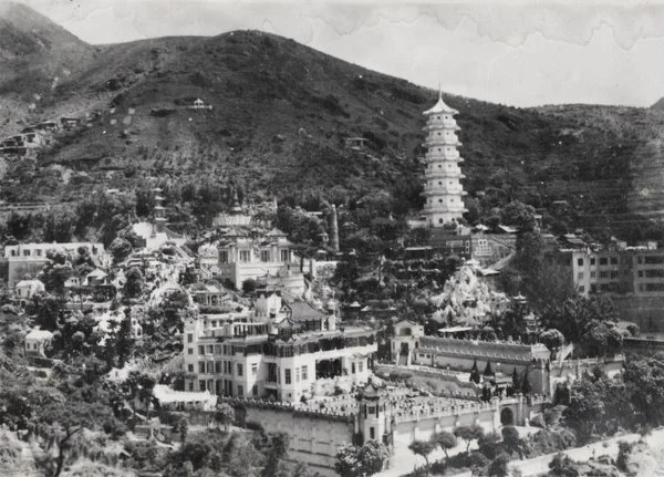 Black and white photograph of a hillside with buildings, a pagoda, and a mountain in the background.