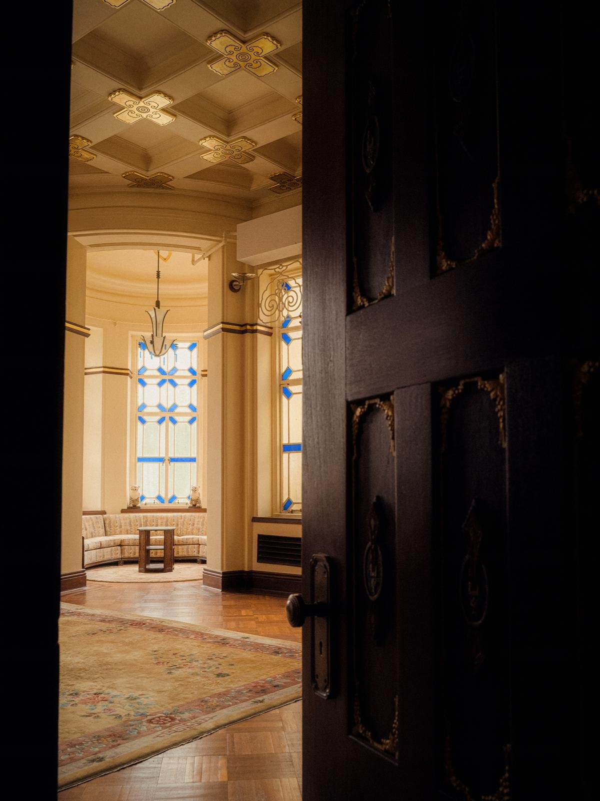 View of an elegant, vintage-style sitting area with large stained glass windows, a curved sofa, a wooden table, and a decorative chandelier, seen through a partially open dark wooden door.