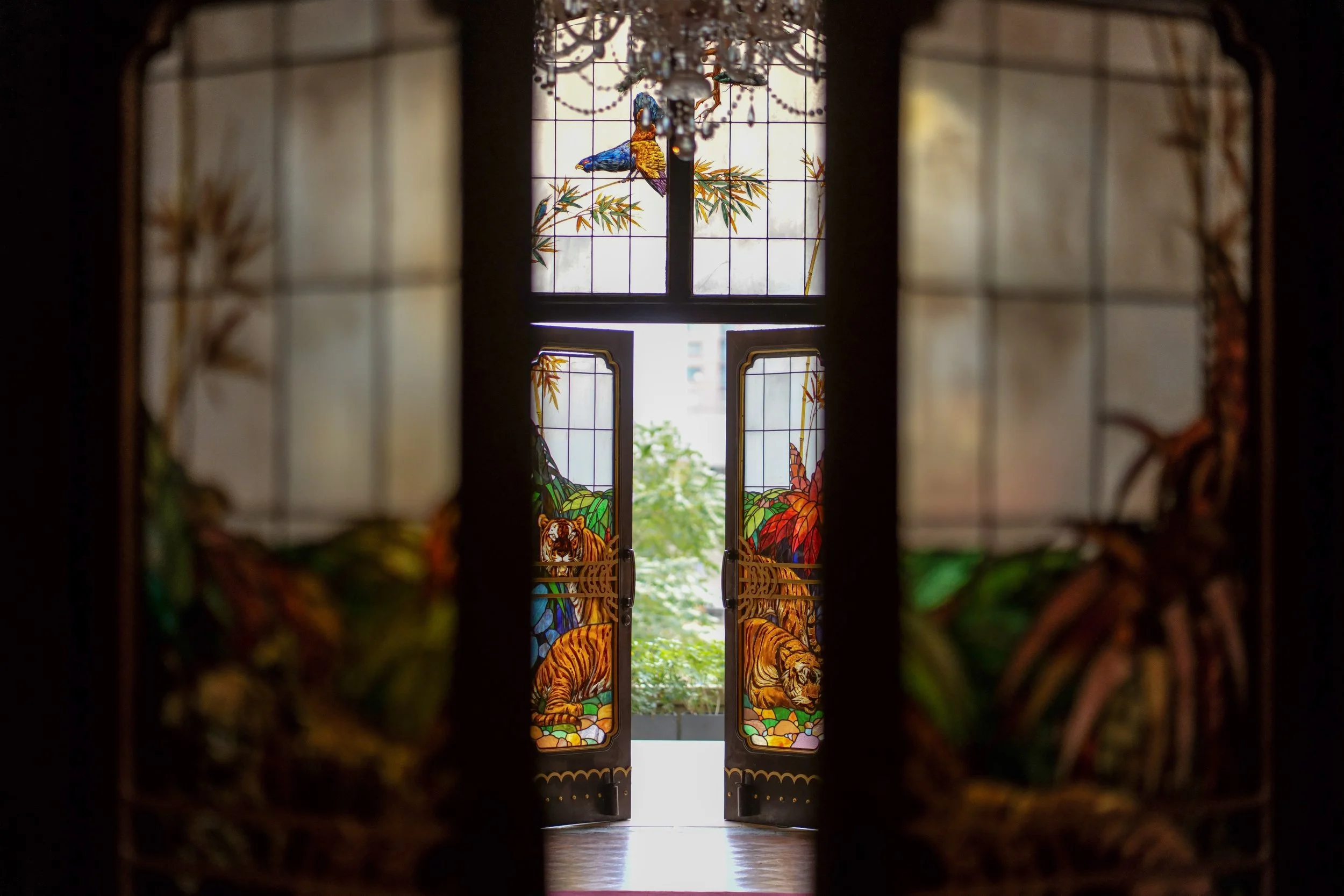 View through a narrow opening of stained glass doors featuring tigers and a lush jungle scene, with the outside visible through the open doors.