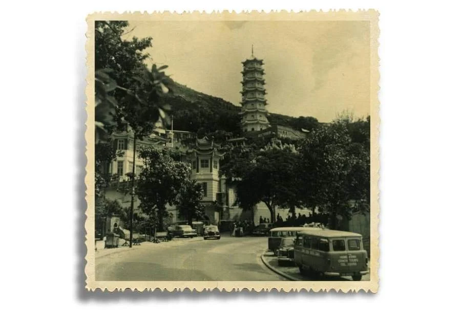 Black and white photo of a street with vintage cars, trees, and buildings, with a multi-story pagoda on a hill in the background.