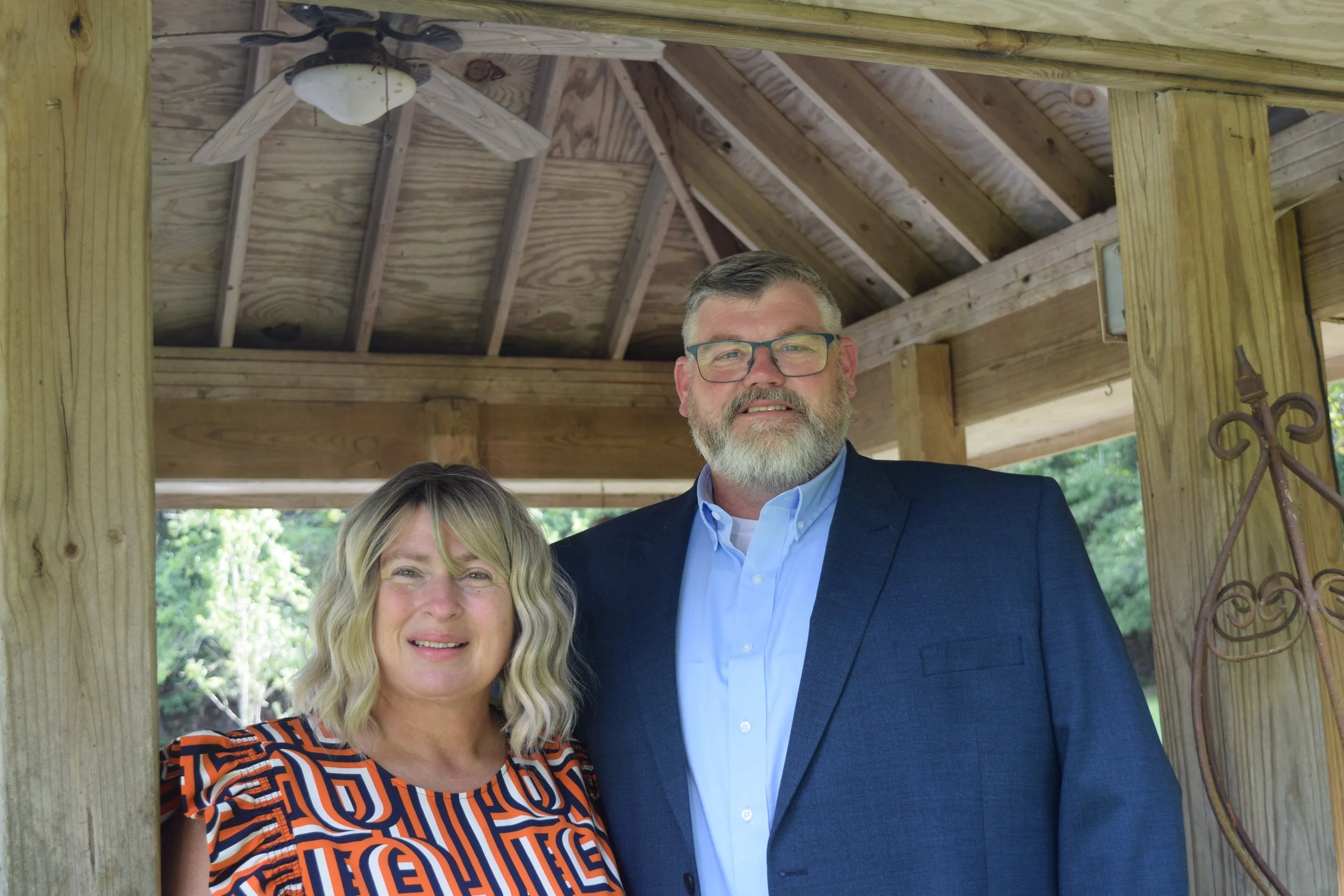 A woman with blonde, wavy hair wearing a colorful geometric patterned top and a man with gray hair, beard, and glasses in a dark blue suit and light blue shirt, standing together under a wooden pavilion with a ceiling fan, surrounded by greenery.