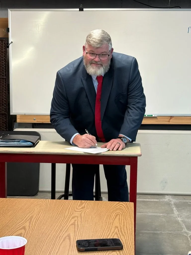 A man with gray hair and a beard, dressed in a dark suit, light blue shirt, and red tie, is standing and signing a piece of paper on a desk in a classroom setting.