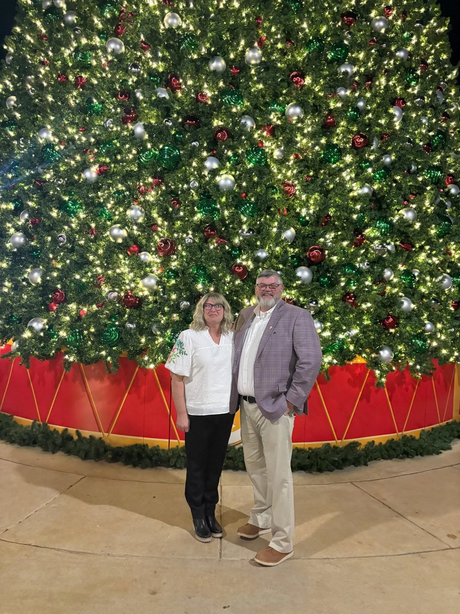 A man and woman standing in front of a large, decorated Christmas tree with lights and ornaments.
