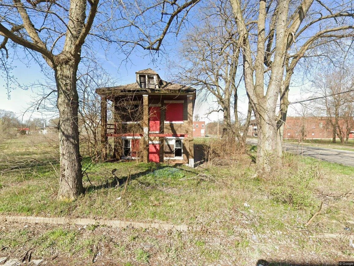 A small abandoned two-story house with boarded-up windows and red paint, surrounded by leafless trees and overgrown grass in an open area.
