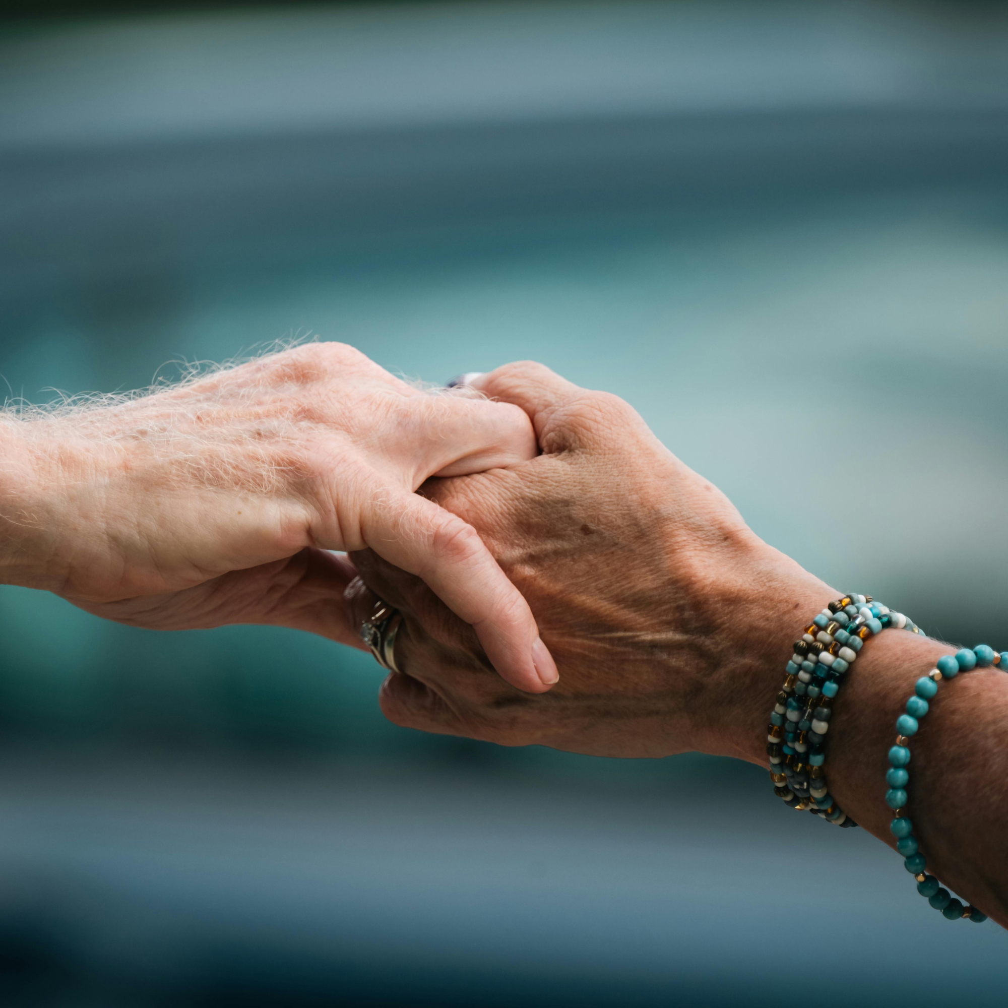 Two hands holding each other, one with a beaded bracelet, against a blurred blue background.