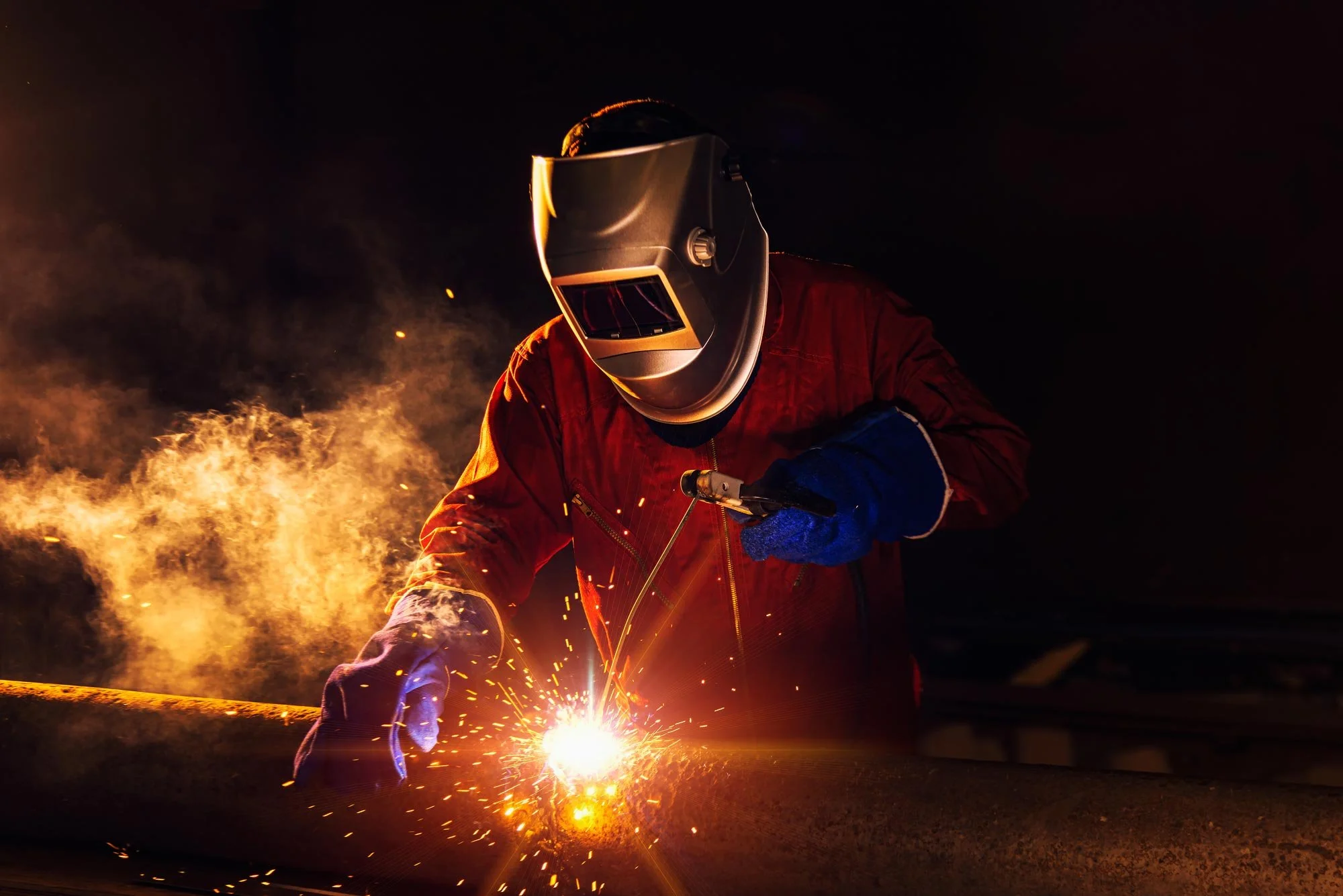 A welder wearing a protective helmet and gloves welding metal at night, with sparks and smoke around.