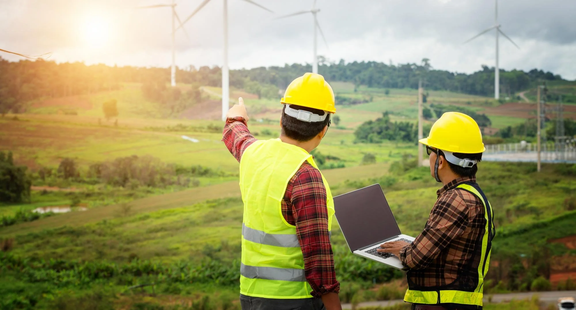 Two construction workers wearing yellow safety helmets and reflective vests using a laptopn and look over a landscape of green fields and wind turbines, with one of them pointing towards the wind turbines.