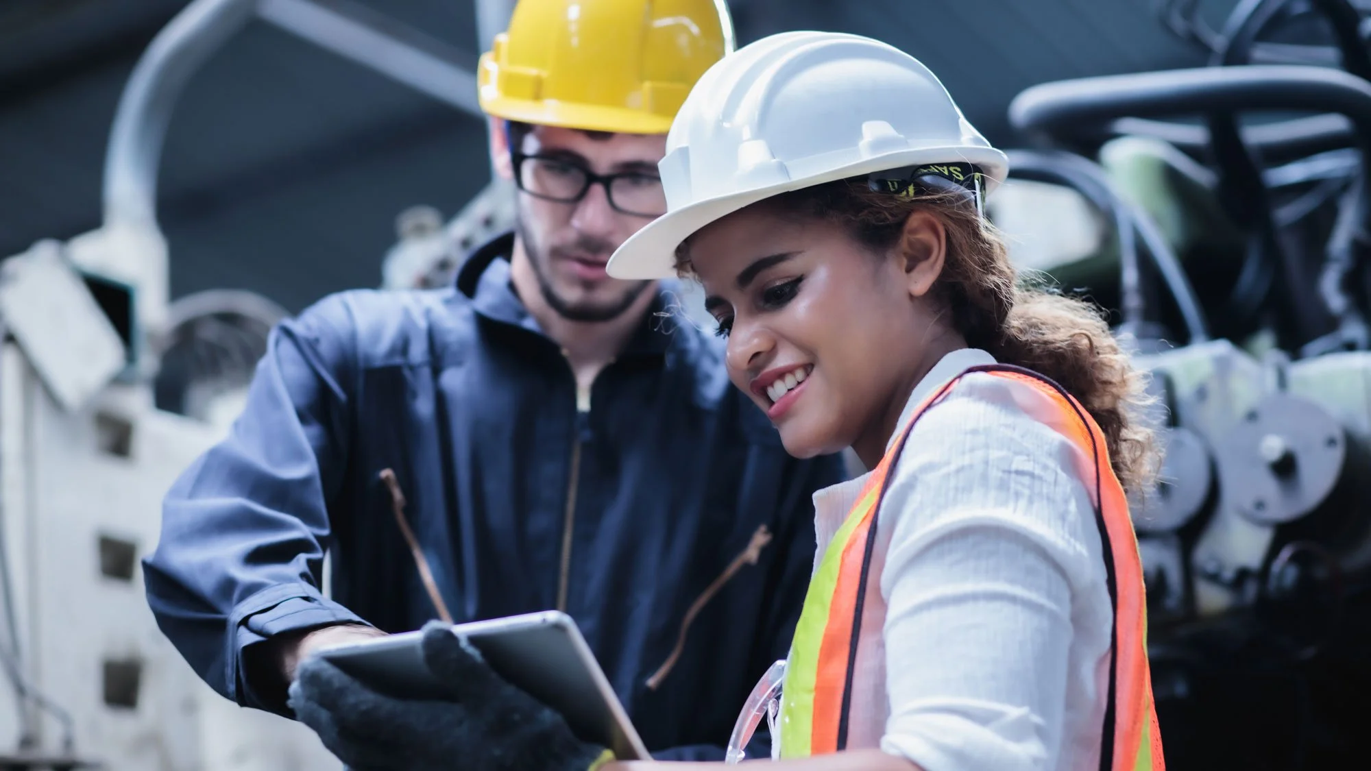 Two workers wearing safety helmets and reflective vests examining a tablet in an industrial setting.