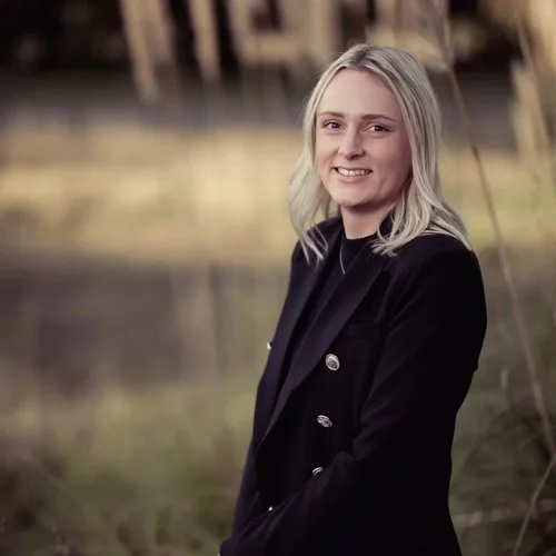 A woman with blonde hair smiling outdoors near a body of water and trees.