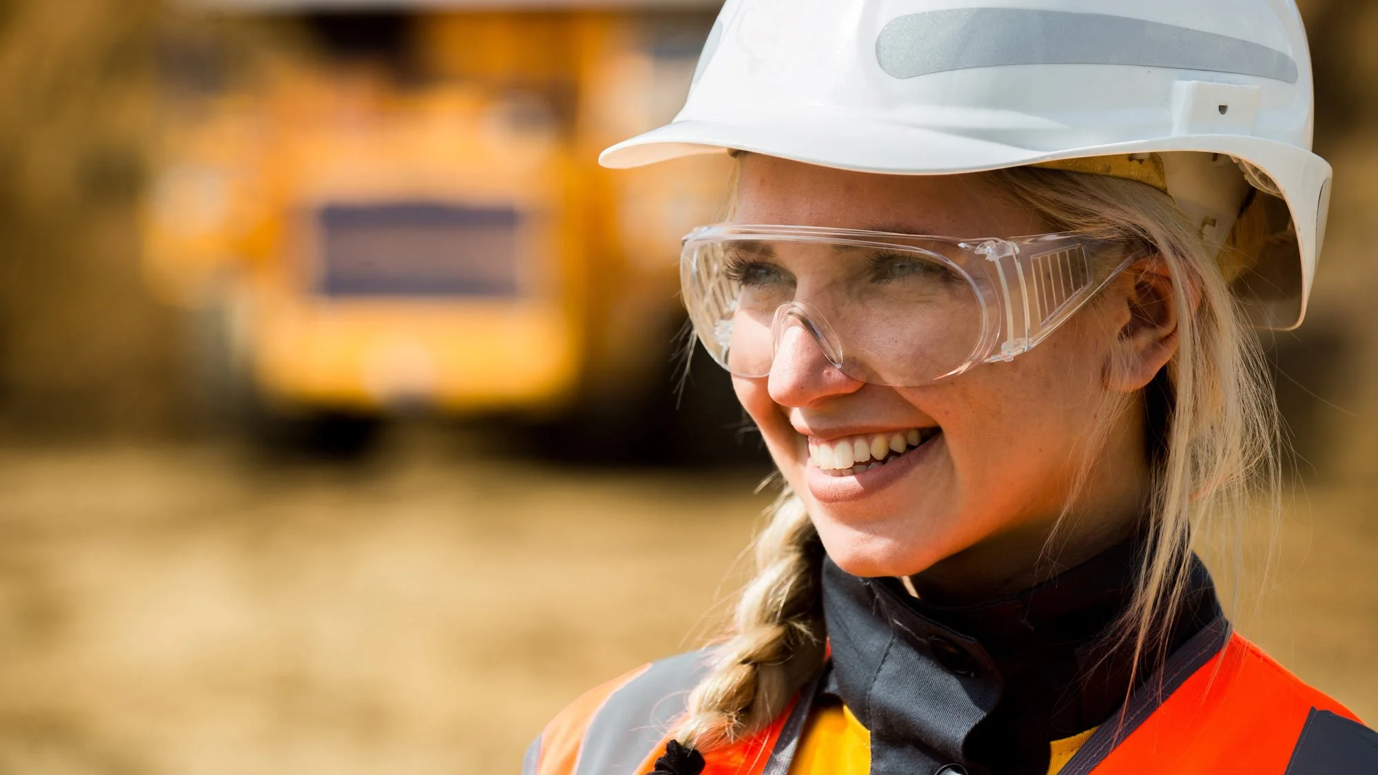 A woman wearing a white safety helmet, clear safety glasses, and an orange safety vest smiling outside with a blurred construction vehicle in the background.