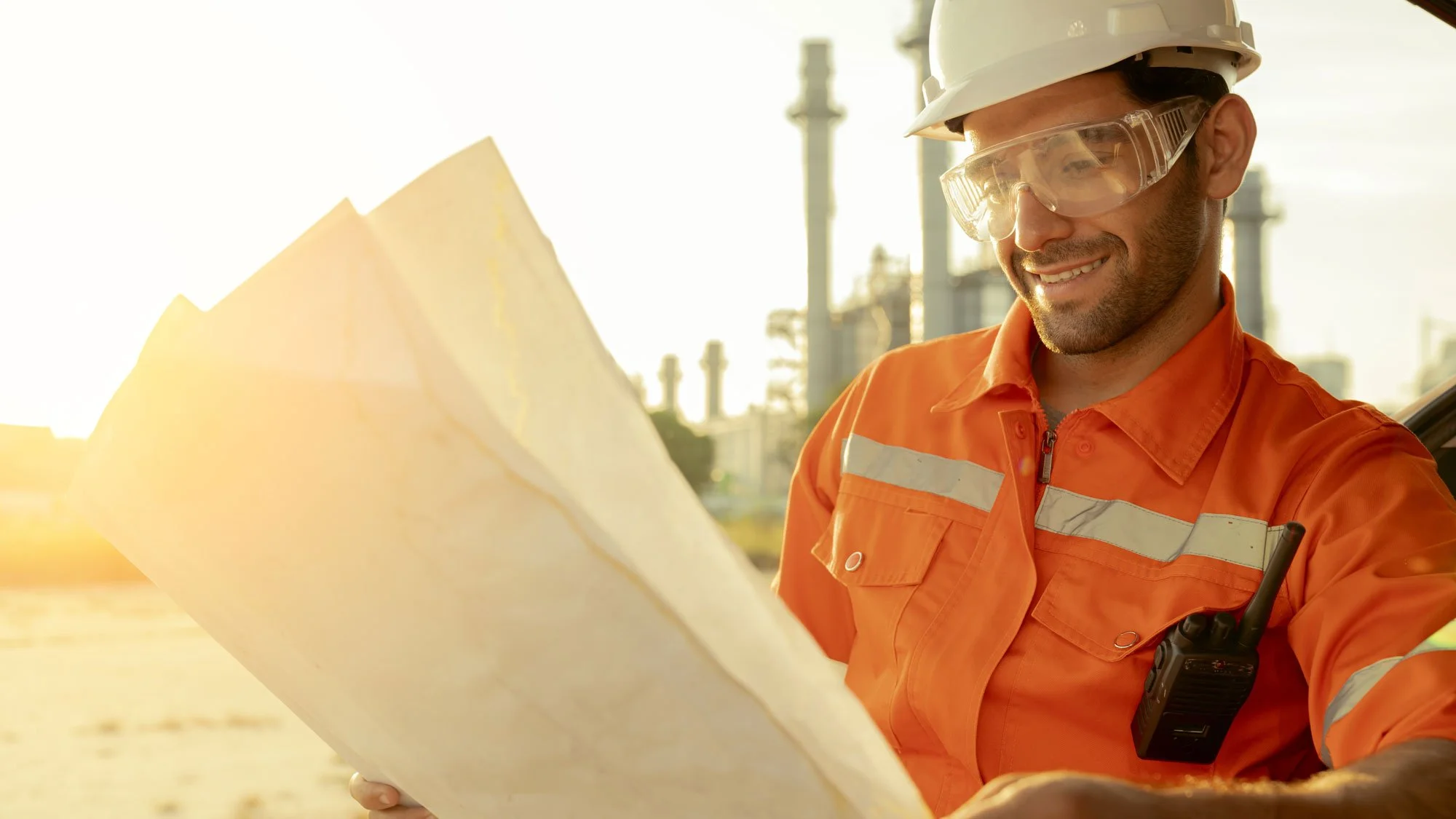 Construction worker in orange safety jacket and hard hat holding and examining blueprints at an industrial site during sunset.