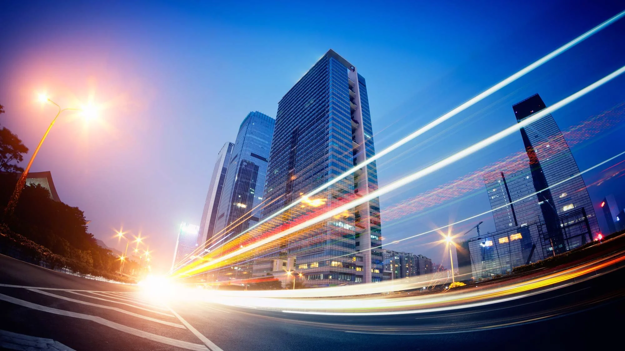Cityscape at dusk featuring tall modern glass skyscrapers and streaks of light from moving vehicles on the road.