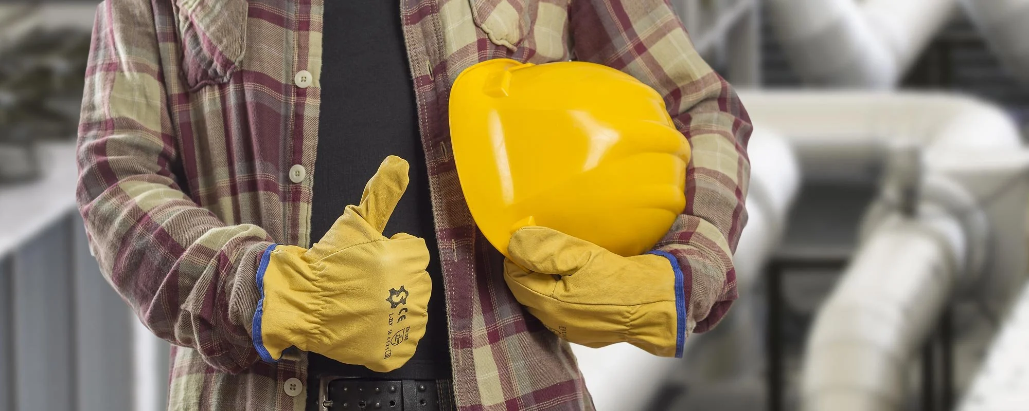 Person wearing work gloves and a plaid shirt holding a yellow hard hat in an industrial or manufacturing setting.