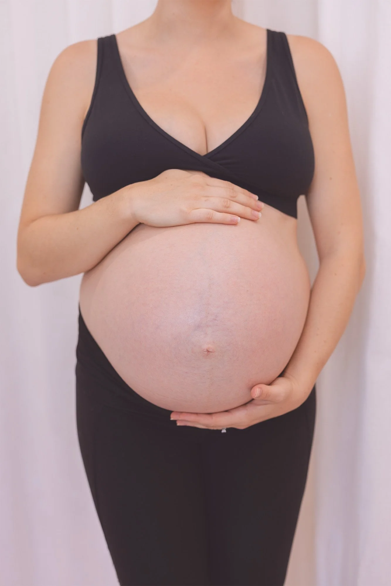 A pregnant woman wearing a black sports bra and black pants, cradling her large belly with both hands, standing against a neutral background.